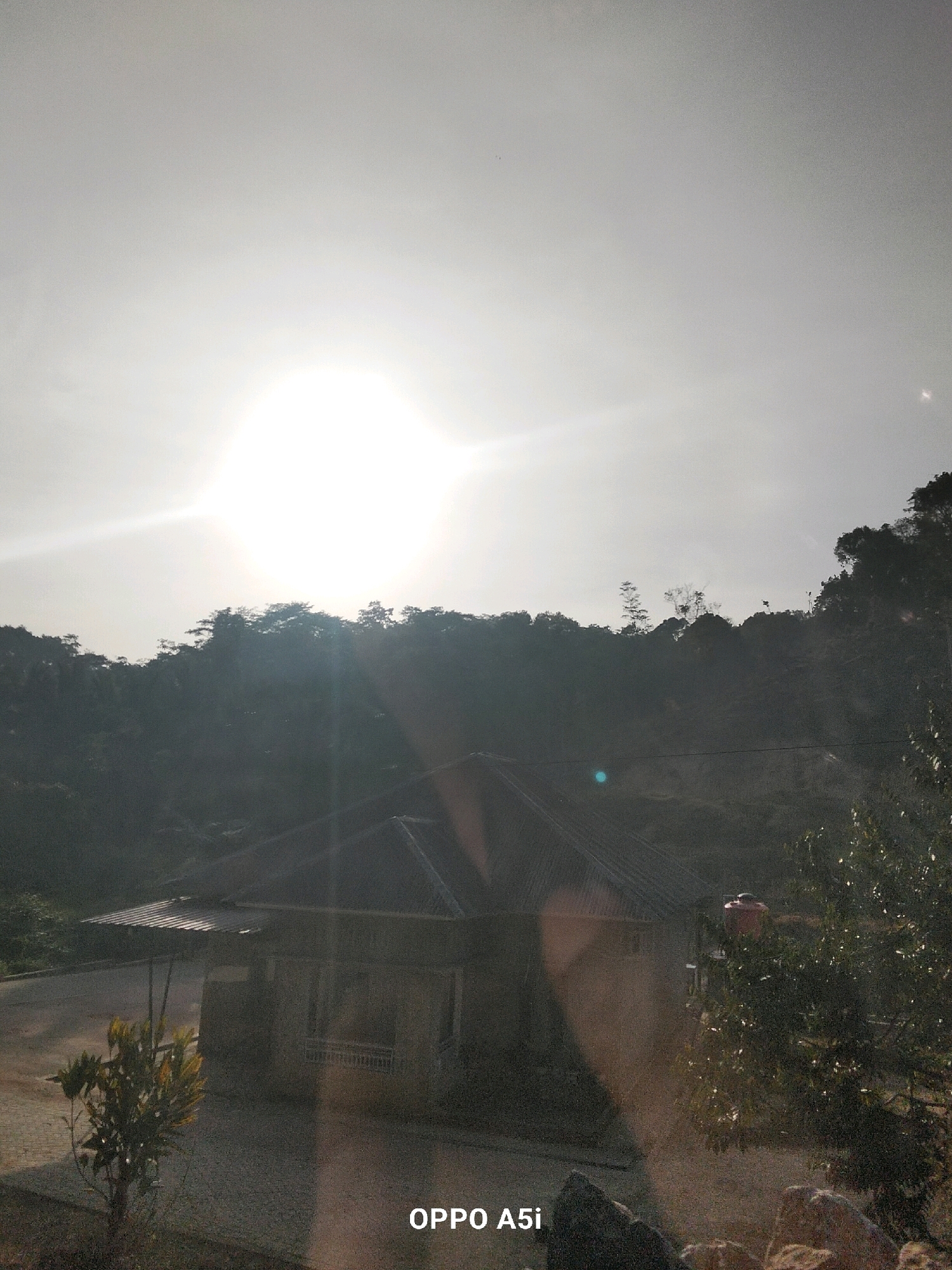 The image captures a sun-drenched scene in Tabanggele, Indonesia. A bright sun dominates the sky, casting a strong glare. In the foreground, a small, single-story building with a dark, pitched roof is visible. The building appears to be made of concrete or stone. Adjacent to it, a small tree with green and yellow leaves stands on a gravelly surface. Further back, a dense, tree-covered hillside rises, silhouetted against the bright sky. A red water tank is perched on the roof of an outbuilding or extension to the main structure. The overall impression is of a rural or semi-rural setting bathed in intense sunlight.