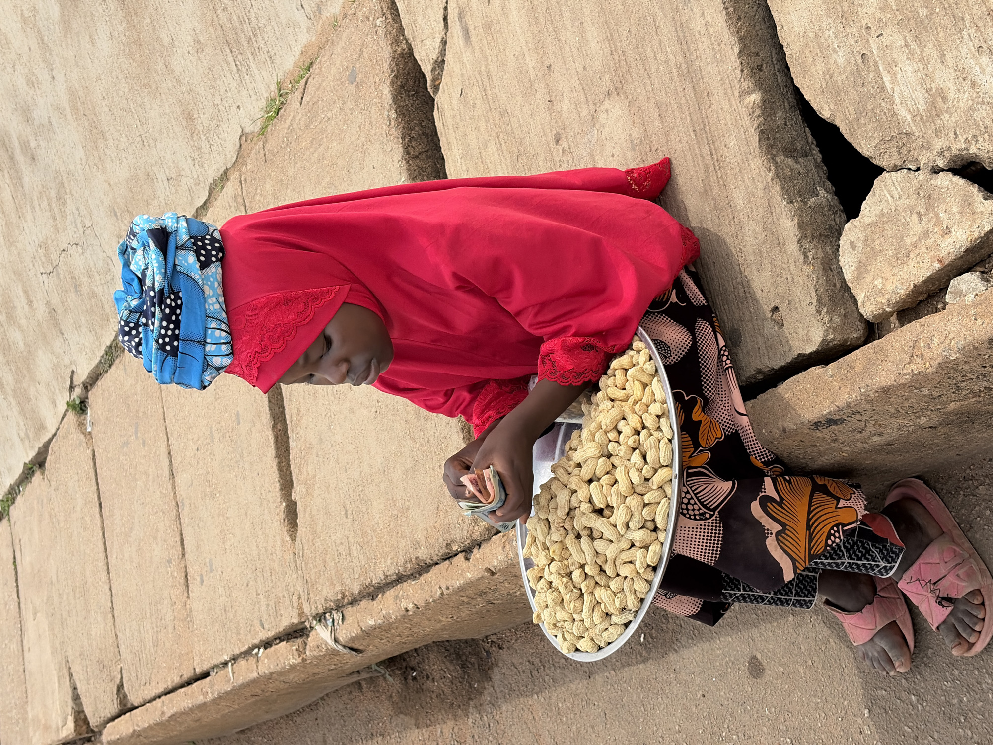 A young girl sits on a concrete structure on a street in Jos, Nigeria, selling peanuts. She wears a vibrant red hijab and a patterned skirt. On her head, she has a blue and black headscarf. Her hands are occupied with a small stack of currency. A large metal tray filled with peanuts sits in her lap. The background shows a concrete sidewalk with cracks and some sparse vegetation. She is wearing pink slip-on sandals.