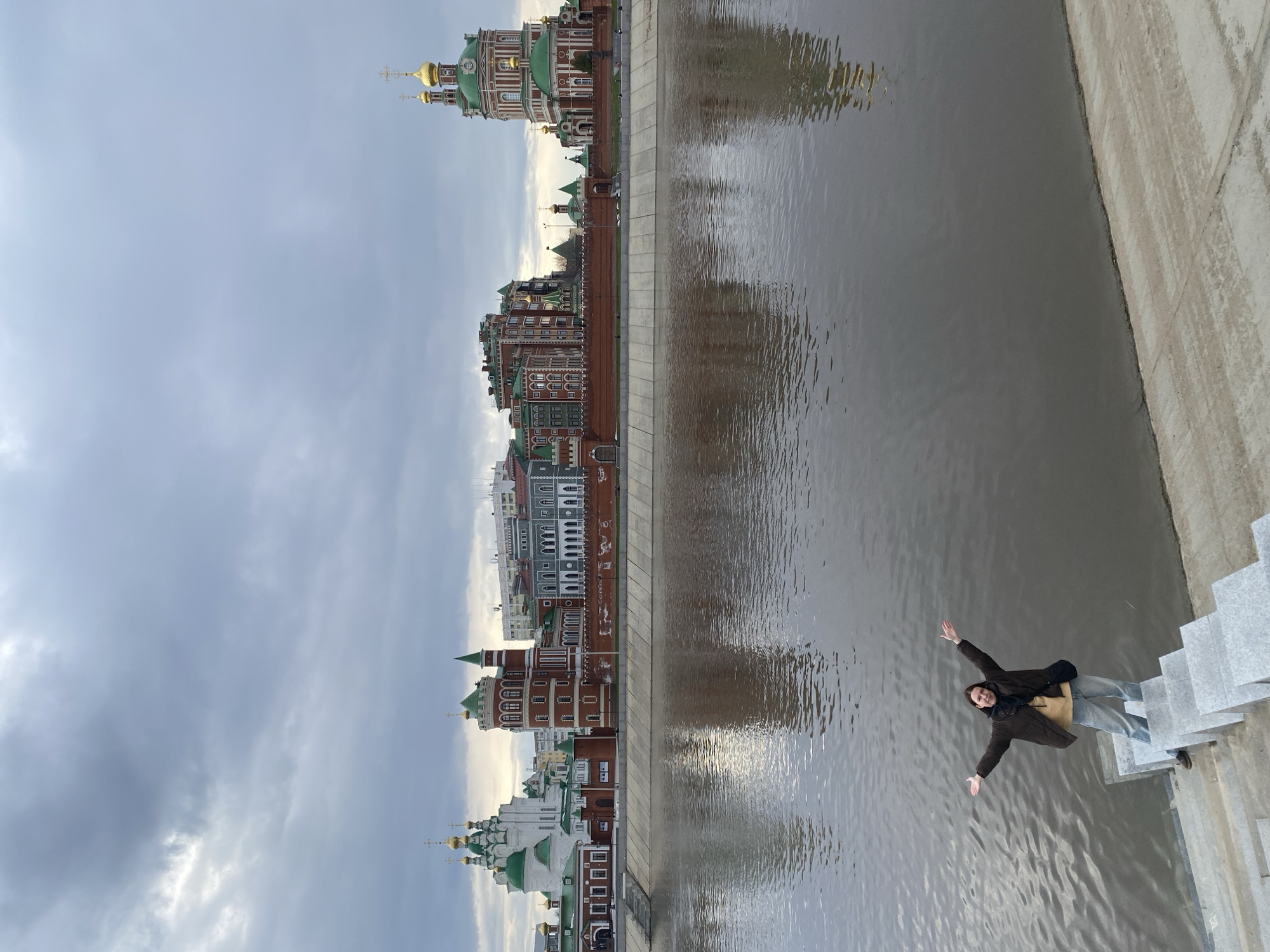 A photograph captures a person posing on a set of light-colored stone steps next to a river, with the cityscape of Yoshkar-Ola, Russia, visible on the opposite bank. The person, appearing to be a young adult, is seen from a slightly elevated angle, smiling cheerfully with both arms outstretched towards the viewer. They are dressed in a dark brown jacket, a lighter-colored shirt underneath, light blue jeans, and dark shoes, with a black bag slung over their shoulder.
The river, a prominent feature, has a grey, somewhat choppy surface, reflecting the overcast sky and indistinct shapes from the buildings. On the far bank, a vibrant and distinctive architectural ensemble lines the river. This includes several buildings with traditional Russian church architecture, featuring green roofs and prominent golden onion domes topped with crosses, alongside multi-story buildings in a mix of red brick, white, and grey facades, adorned with decorative elements and pitched roofs. A sturdy concrete or stone embankment runs along both sides of the river.
The sky is uniformly overcast, indicating daytime but without direct sunlight. The overall atmosphere appears calm and pleasant, with the person's pose conveying happiness and relaxation during a visit to the picturesque location. No discernible text is visible within the image.