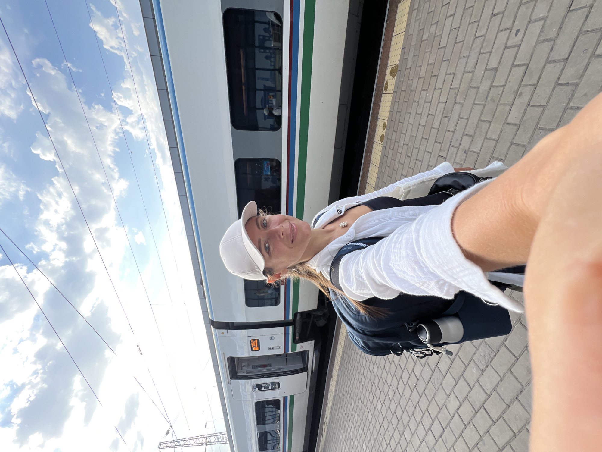 In Tashkent, Uzbekistan, a woman stands on a paved train platform, taking a selfie in front of a modern white train with blue and green stripes. She wears a white baseball cap, a white open shirt over a black top, and a necklace with a pearl pendant. A dark backpack with a silver water bottle in a side pocket is slung over her shoulders. The train doors are open, revealing passengers inside. Overhead, power lines crisscross against a bright sky with scattered clouds. The platform is constructed of interlocking gray bricks with yellow tactile paving near the train.