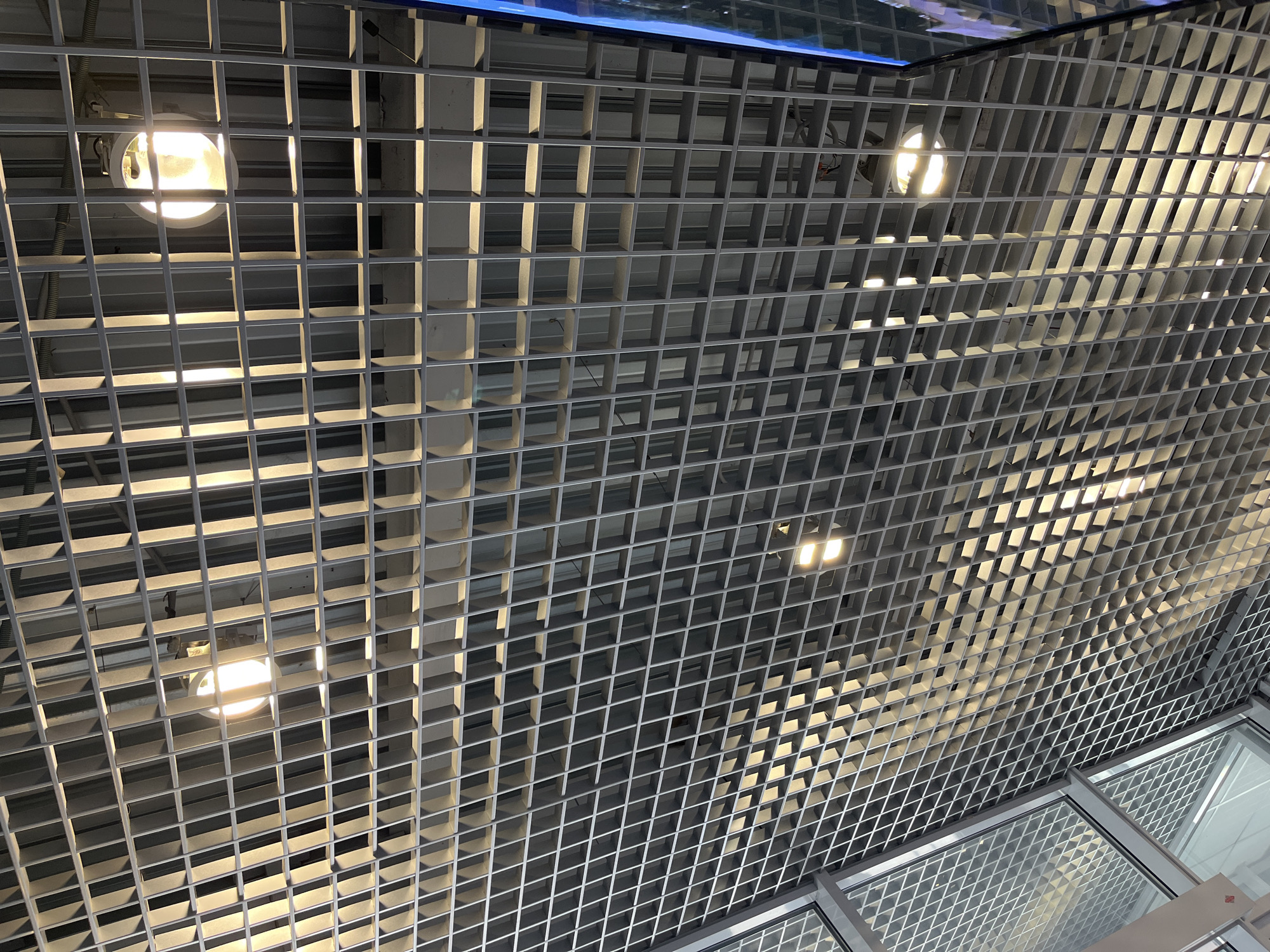 The image shows a ceiling consisting of a dense, metallic grid pattern. Embedded within the grid are several light fixtures, emitting a warm, yellow light. The grid is illuminated by the lights and ambient light, creating varying shades of grey. A section of glass paneling is visible at the bottom right corner of the image. The location is not discernible from the image alone. There are no people, subjects, objects, actions, or interactions visible other than the elements described above. The environment is an indoor space with an industrial or modern architectural style.