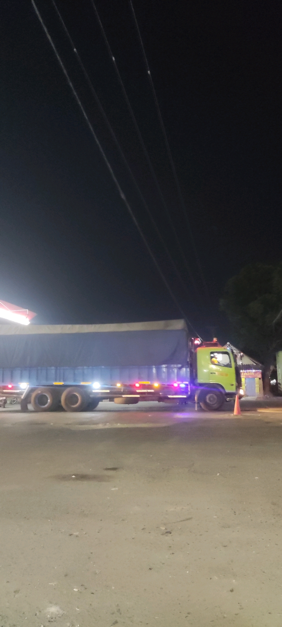 The image shows a semi-trailer truck parked at night on a paved surface, possibly a road or parking lot. The truck has a green cab and a long, dark blue tarp-covered trailer. Various lights are visible on the truck, including headlights, taillights, and what appear to be marker lights along the side. There's a traffic cone placed near the front wheel of the truck. In the background, there are some buildings with lights and trees. The sky is dark, indicating it is nighttime. There are visible power lines running diagonally across the upper portion of the image. The ground appears to be a mix of asphalt and dirt.