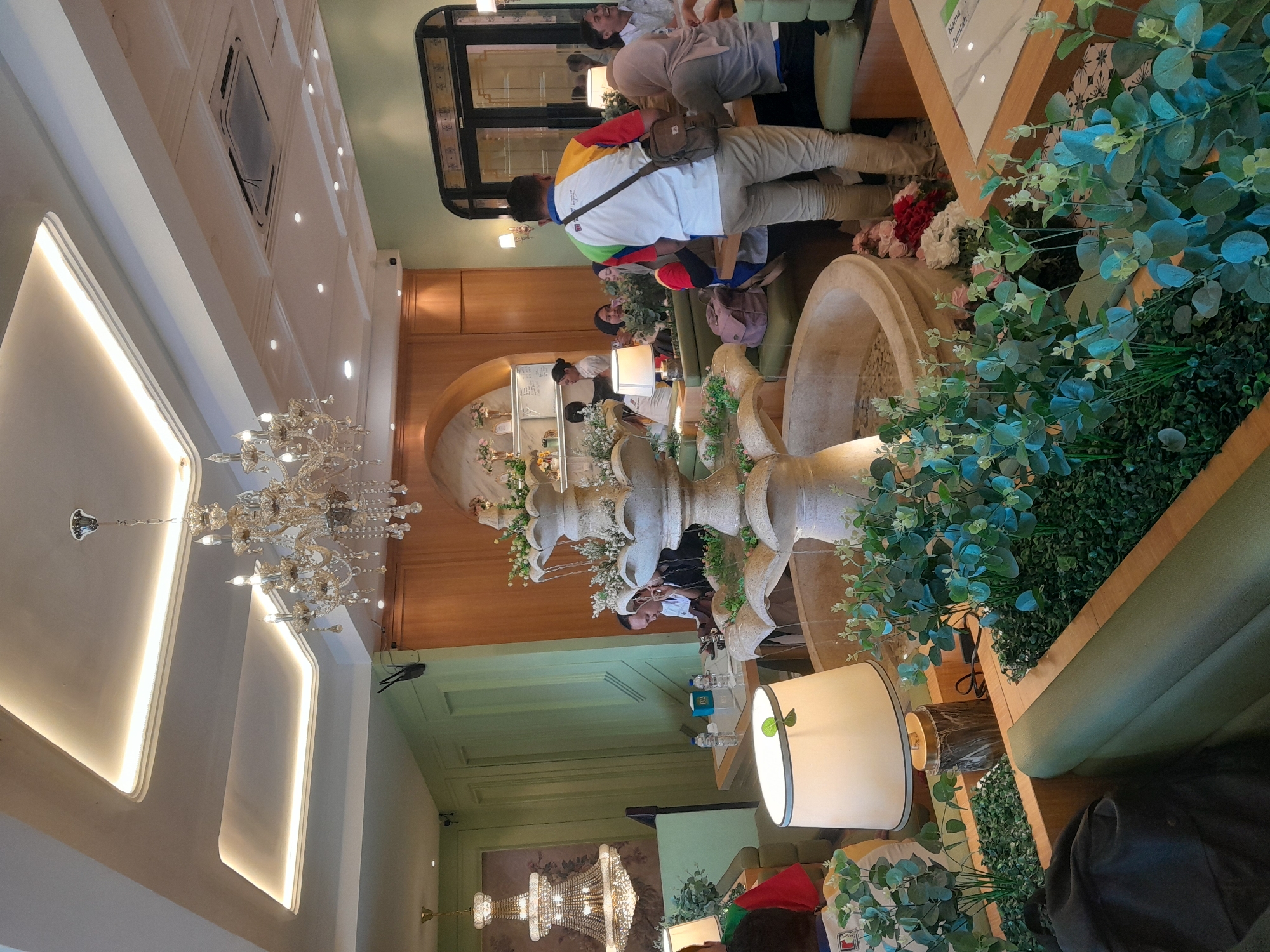 The image captures an indoor scene, likely a cafe or restaurant, with a decorative water fountain as a central focal point. The fountain is made of stone and features multiple tiers with water cascading down. It is surrounded by lush green plants and artificial foliage, creating a vibrant, naturalistic atmosphere.
The interior design appears to be elegant and modern, with white paneled ceilings that incorporate recessed lighting. A large, ornate crystal chandelier hangs prominently from the ceiling, adding a touch of grandeur. Other lighting fixtures include lamps with white shades placed strategically on tables.
Several people are visible in the scene, engaged in various activities. Some are seated at tables, suggesting they are patrons. One man, wearing a colorful shirt, is standing and looking towards the right. Another person is seen from the back, with a bag slung over their shoulder. The overall ambiance suggests a lively but relaxed environment.
The background features wooden paneling and light green walls, contributing to the pleasant and inviting decor. In the far left background, another elaborate chandelier can be seen, hinting at a larger establishment. The flooring appears to be tiled. The image is taken from a slightly elevated perspective, looking down towards the fountain and surrounding seating area.
While the image depicts an interior, the prompt specifies the location as the city of Sempusari, Indonesia. This information suggests the cafe is situated within this Indonesian city.