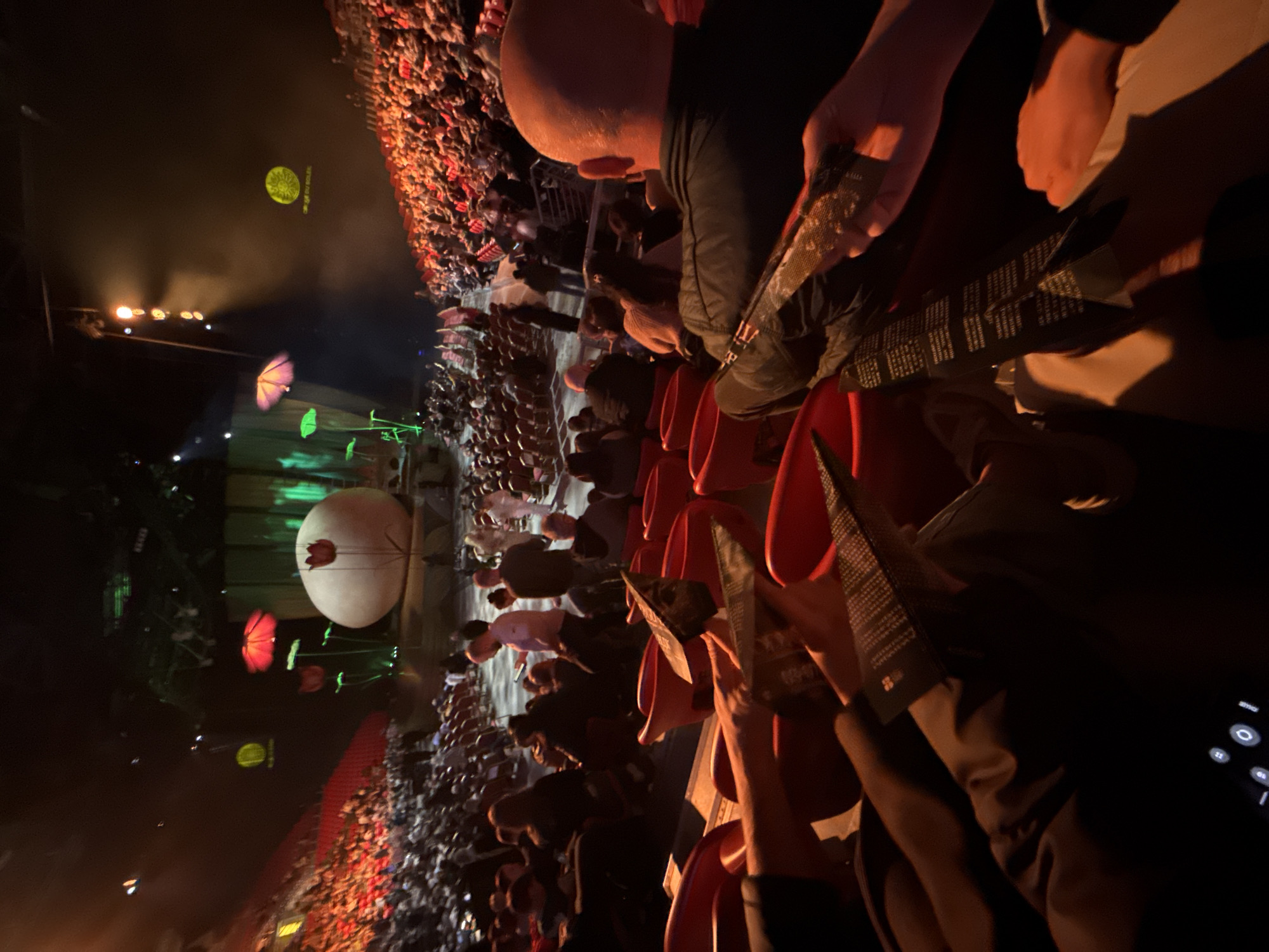 The image captures a live event with a large crowd, likely a concert, at night. The perspective is from a seated position in the audience. A stage is visible in the distance featuring large illuminated objects resembling a white sphere and colorful flower-like structures. Above the stage is a green circular logo and some lights. The crowd fills the space between the viewer and the stage. Many people are holding up red objects. The person taking the photo is holding a leaflet. The photo was taken in Sofia, Bulgaria.