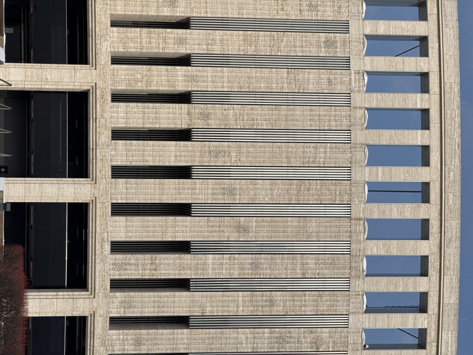 This image displays the facade of a multi-story building located in Plodorodny, Russia. The uppermost visible section features a row of nine tall, narrow, rectangular windows, through which a clear blue sky is visible. White accumulations, consistent with snow, rest on the ledges directly beneath these windows. The central facade consists of light-colored, vertically textured stone, possibly travertine. This section is characterized by a series of evenly spaced vertical elements, each comprising multiple narrow, dark vertical stripes or louvers. Beneath these striped sections are deeper, recessed vertical openings, appearing dark. Between these dominant vertical features, subtle, repeating decorative patterns are embossed or carved into the stone surface, resembling stylized floral or geometric motifs. The lowest visible portion of the building showcases larger, recessed rectangular openings framed by broad, light-colored stone, with dark glass or panels filling these apertures, likely indicating ground-floor entrances or large windows. A small area of reddish-brown vegetation is visible in the bottom right foreground. No individuals are present, and no actions or interactions are depicted within the frame. The scene is illuminated by natural light, suggesting daytime.