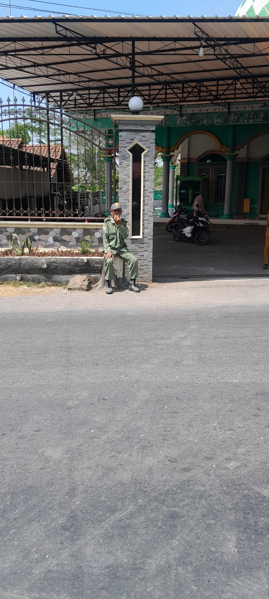 In Jeruklegi Kulon, Indonesia, a man in a green uniform sits on a log by the roadside. He wears a matching hat and dark boots. The man is facing left, with his right hand near his mouth, possibly holding a cigarette. Behind him, a modern building with green accents and a white roof is visible, identified by signage as "MASJID AL MAFAATIH". A fence with a decorative metal gate is to the left of the man. Motorcycles are parked in front of the building. The ground is paved with asphalt, and the weather appears sunny.
