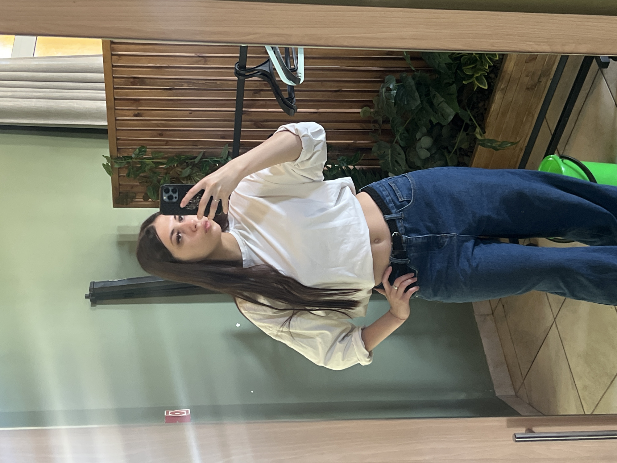 A young woman with long, dark hair is taking a mirror selfie. She is wearing a white, cropped, long-sleeved shirt and dark blue, high-waisted jeans. Her left hand is on her hip, and her right hand is holding a black smartphone. She is standing in front of a mirror, which reflects the scene. The background shows a light green wall, a wooden slatted panel with plants, and a wooden closet or door. A green bucket is visible on the floor. The image appears to be taken indoors.