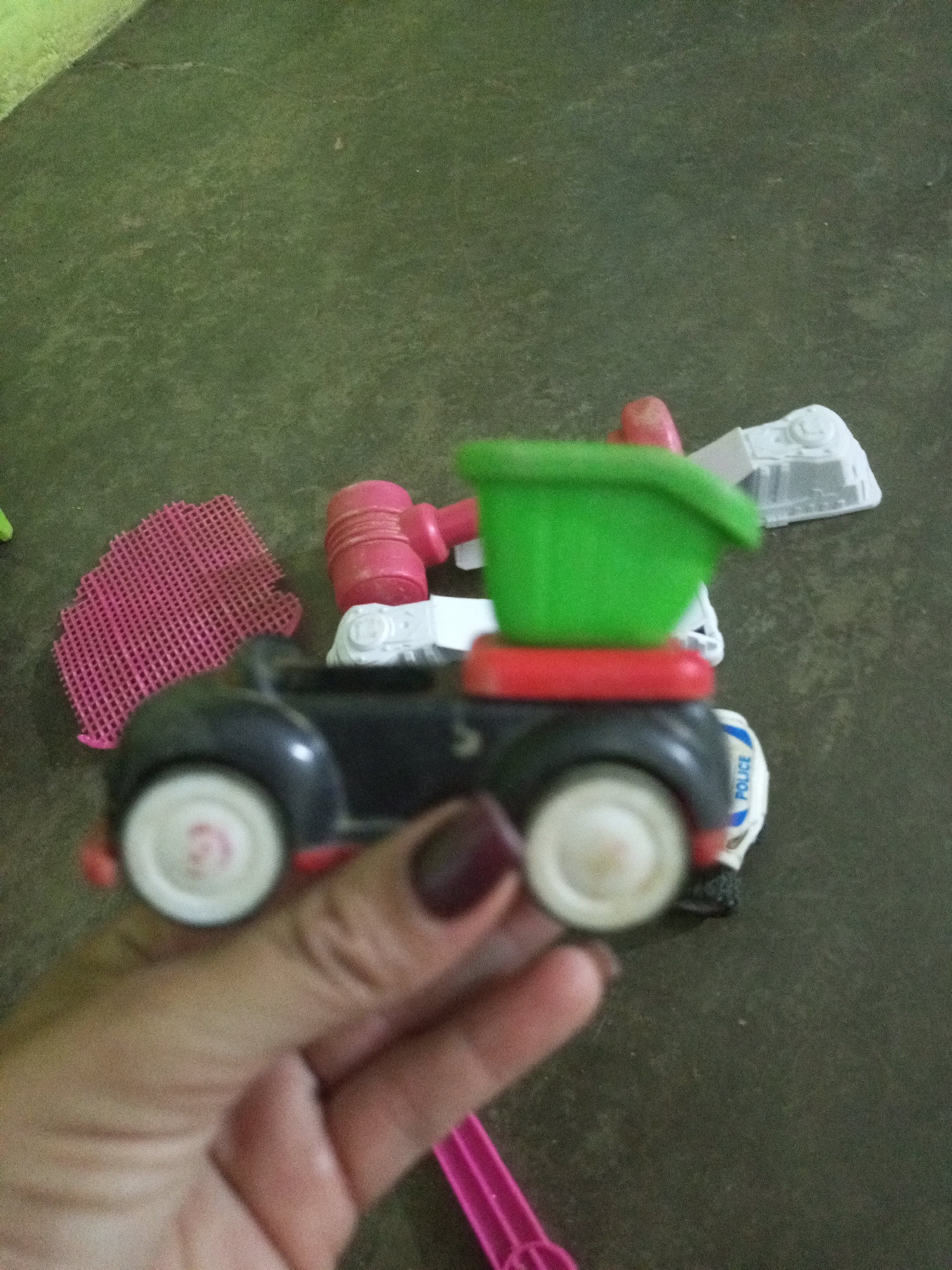 The image shows a close-up of a toy car with a green bucket on top, held by a person's hand. The car is black with red trim and white wheels. There are other toys in the background, including a pink mesh object and a pink hammer. The scene appears to be indoors, on a textured, dark-colored surface. The location is identified as La Tagua, Venezuela.