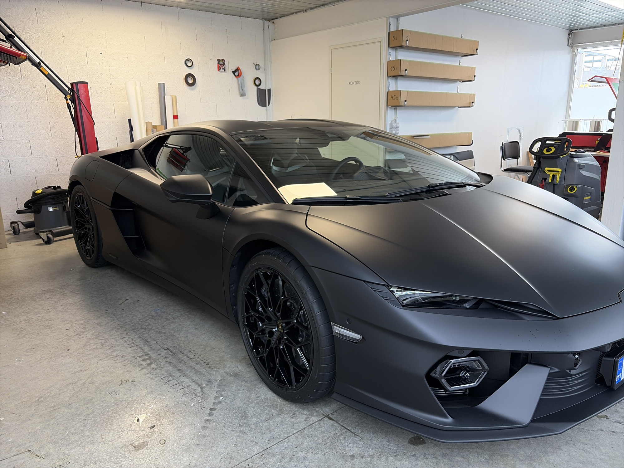 A matte black Lamborghini is parked inside a garage in Peetri alevik, Estonia. The car is the primary subject, positioned in the center of the frame, angled towards the right. Its sleek, dark exterior contrasts with the surrounding environment. The garage appears to be a workshop or storage space, with a concrete floor marked by tire tracks and general wear. The walls are painted white, and various items are visible, including rolls of material on the left, possibly for car wrapping or window tinting. On the back wall, there are shelves with cardboard boxes, a door labeled "KONTOR" (Office), and a chair. To the right of the car, a piece of industrial cleaning equipment, possibly a large vacuum cleaner or scrubber, is present, along with a red and black cart. The lighting in the garage is bright, suggesting artificial overhead lights. No people are visible in the image.
