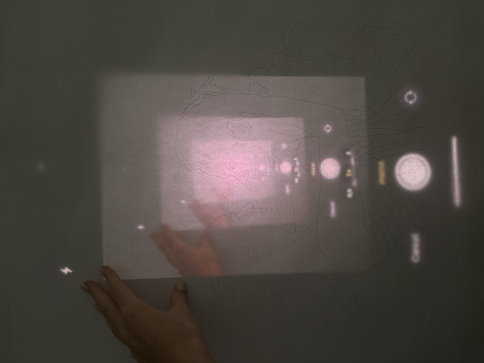 The image shows a hand holding a semi-transparent paper against a light gray surface, possibly a wall or large paper. The paper appears to have an underlying pink projection or light shining through it. A faint pencil sketch of a face and flowing hair is visible on the surface, partly obscured by the paper. There are various out-of-focus light orbs and streaks projected onto the surface around the sketch. The photo does not provide enough information to determine if it's located in Choeng Thale, Thailand.