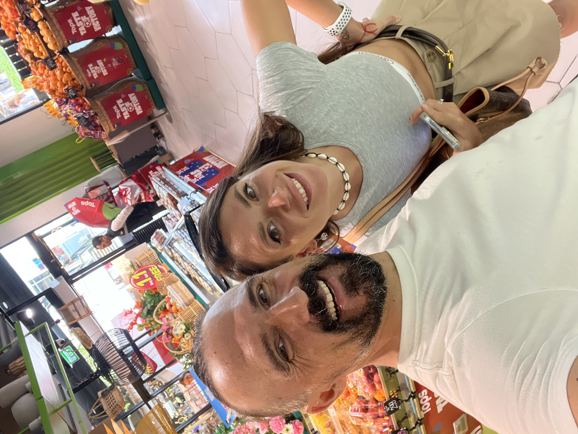 A man and a woman are taking a selfie in what appears to be a supermarket. The man is in the foreground, smiling broadly, with a beard and short hair. He is wearing a white t-shirt. The woman is to his right, also smiling, with long dark hair. She is wearing a grey crop top and a beige skirt. She has a shell necklace and a watch on her left wrist. She is holding a smartphone in her right hand. In the background, there are displays of fruit and promotional materials, with the text "Tops Taste of Victory" visible. The environment suggests a bright, indoor retail space. The location provided is Baan Chaweng, Thailand, which is a tourist area on the island of Koh Samui, known for its beaches and shops.