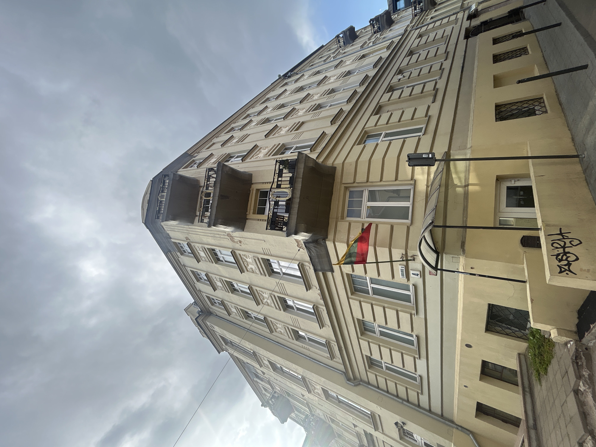 A low-angle, wide shot captures the exterior of a cream-colored, multi-story building on a cloudy day in Vilnius, Lithuania. The building, which appears to be an apartment or office building, features numerous rectangular windows and decorative architectural elements above them. Several balconies with black metal railings jut out from the upper floors. A Lithuanian flag is prominently displayed on a pole near the corner of the building, at the intersection of Labdarių and Tilto streets, indicated by street signs. The ground floor has a covered entrance with a transparent awning and a glass door. Some windows on the lower level are barred. The sidewalk in front of the building is paved with grey tiles, and a low concrete wall with graffiti lines the foreground. The sky is overcast with dramatic clouds.