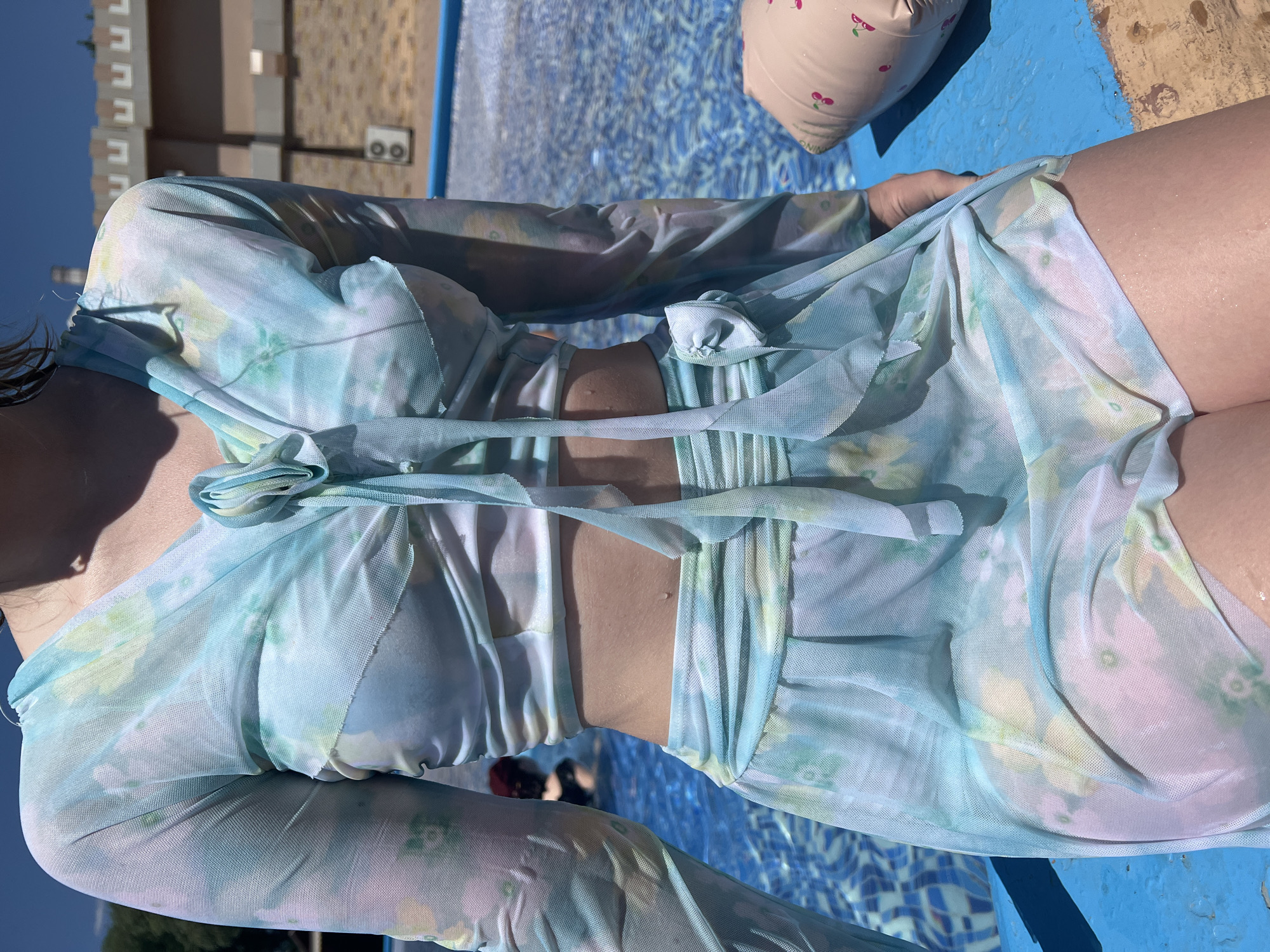 A person is sitting by a swimming pool, wearing a sheer, long-sleeved, floral-print crop top with a tie-front and matching drawstring skirt. The person's midriff is exposed. The swimming pool is blue with a tiled bottom. In the background, a building with a textured facade and two outdoor air conditioning units is visible. The person is sitting on a blue ledge or platform next to the pool. A pink inflatable object with a cherry pattern is partially visible to the right of the person. The sun is shining, casting shadows. The location is a swimming pool area in Shymkent, Kazakhstan.