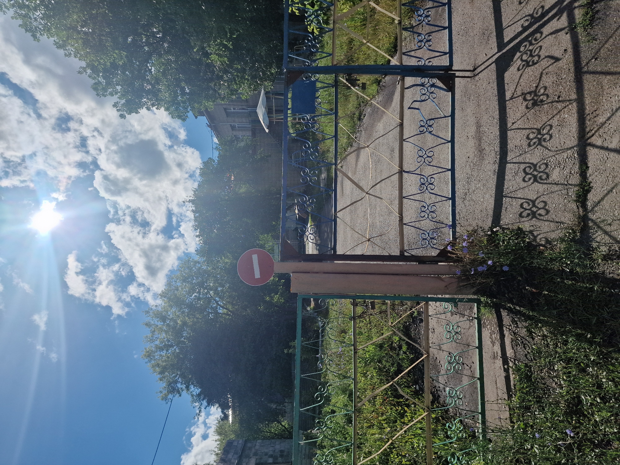 The image depicts a sunny day in Kashgar City, China. The foreground is dominated by a decorative metal gate with ornate scrollwork, painted in blue and yellow hues. A red circular road sign with a white horizontal bar, indicating "no entry," is attached to a wooden post beside the gate. Beyond the gate, lush green vegetation and trees line a paved path. In the background, a building with a pitched roof and windows is partially visible, surrounded by trees. The sky is a vibrant blue, dotted with fluffy white clouds, and the sun is shining brightly, casting long shadows on the ground, suggesting it is daytime. The overall scene evokes a sense of a quiet, possibly residential or public area, with the presence of the road sign indicating some form of access restriction. There is no discernible activity or people in the image.