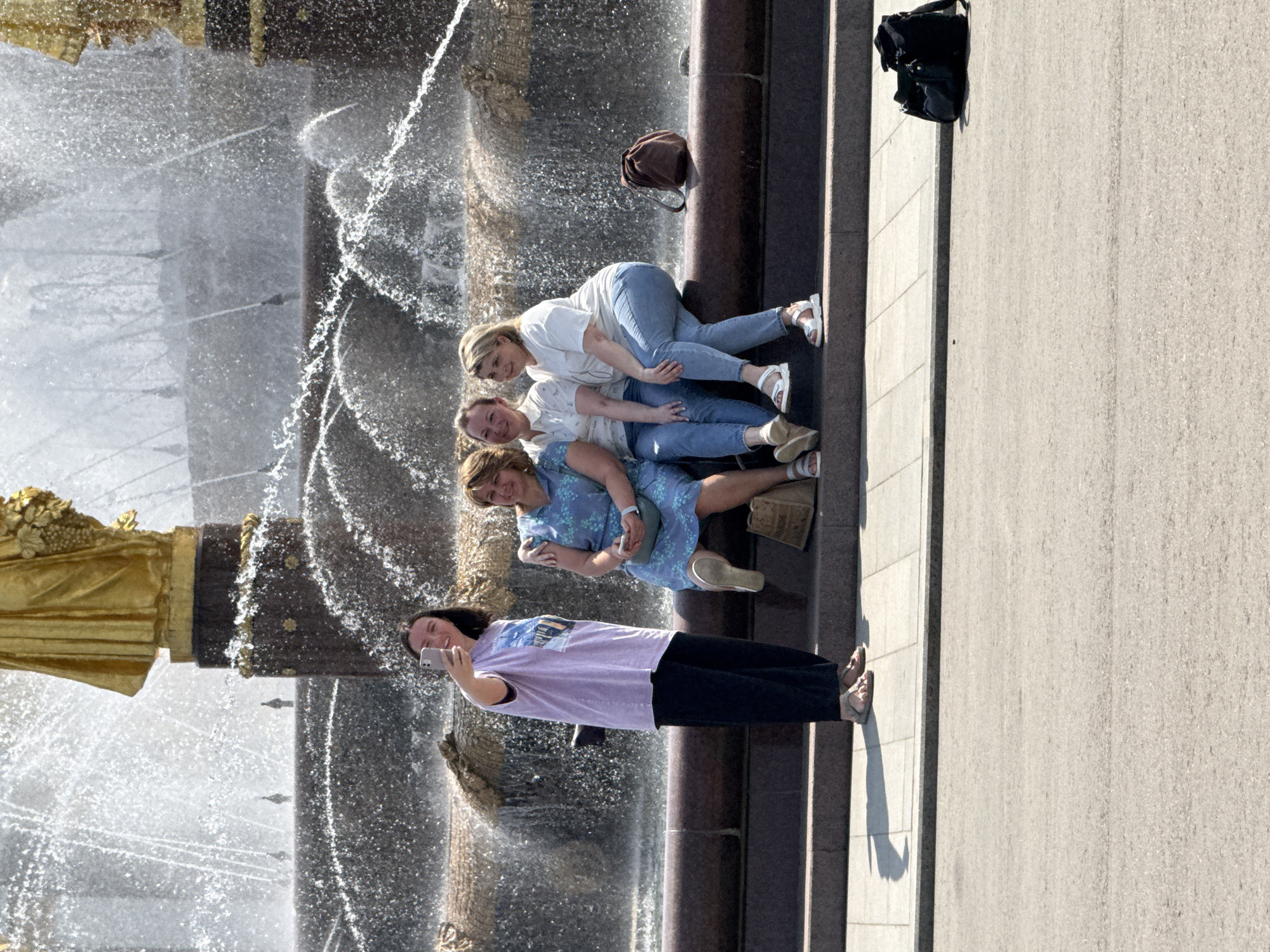 A photograph captures a group of four women posing together next to the Friendship of Nations fountain in Moscow, Russia. The fountain is visible in the background, with water cascading around a golden statue. One woman, closest to the camera, is holding a smartphone and appears to be taking a selfie of the group. The other three women are partially lying on the edge of the fountain structure, arranged one on top of another. A couple of bags are also visible on the ledge of the fountain.