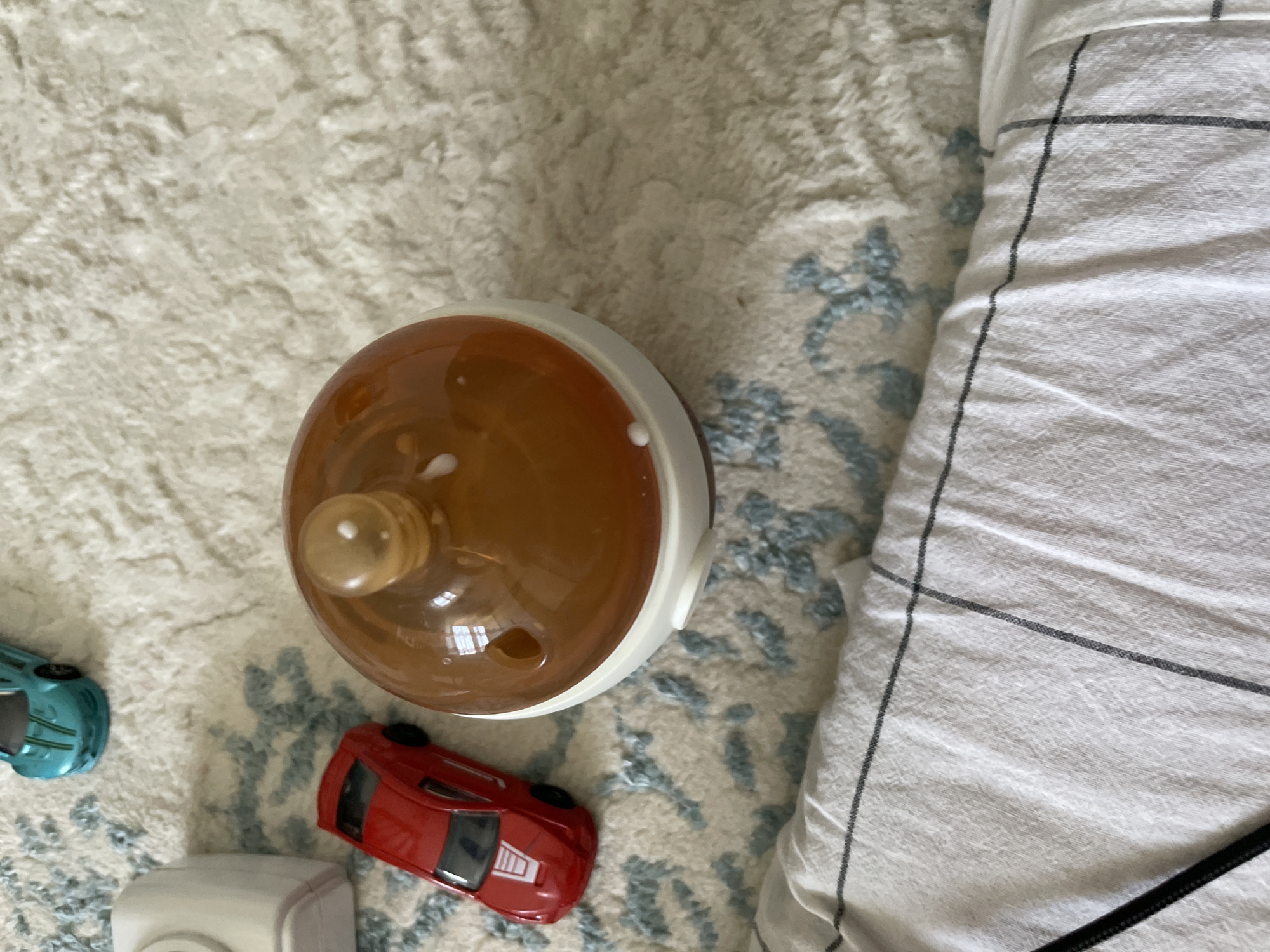 This overhead, close-up shot captures a flat lay arrangement of various items on a textured, off-white rug with a subtle blue floral pattern. The primary subject is a wide-mouthed baby bottle, its amber-colored glass body partially filled with a light brown liquid. A translucent, tan-colored nipple is visible at the top, with a few small white specks, possibly milk residue, clinging to its surface and the rim of the bottle.
To the left of the bottle, a small, teal-colored toy car is partially visible, its metallic finish reflecting the light. Below and to the right of the bottle sits a small, bright red toy car, also with a metallic sheen. In the bottom left corner of the frame, a white object, possibly a baby monitor or a small electronic device, peeks into view. To the right of the scene, a neatly folded white duvet or blanket with thin, dark gray checkered stripes takes up a significant portion of the frame, creating a strong linear element.
The lighting suggests it's daytime, with soft light illuminating the scene. The overall impression is that of a nursery or a child's play area, with the toys and baby bottle suggesting the presence of an infant. The image does not contain any visible text. The location context of Чат-Көл, Kyrgyzstan, is provided as external information and is not directly discernible from the visual content of the image itself.