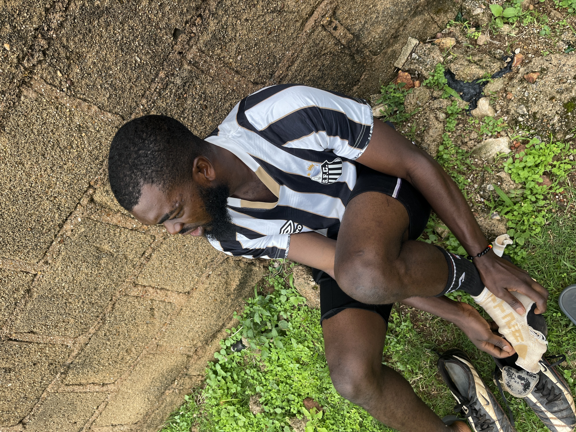The media file shows a person sitting on the ground, adjusting a sock on their foot. The person is wearing a black and white striped jersey with a crest on the chest that reads "S.E.C.". They are also wearing black shorts and black socks with white stripes. The ground appears to be a mixture of dirt, grass, and some broken concrete. There are also a pair of soccer cleats next to the person. The scene suggests an outdoor setting, possibly a field or a yard, with natural lighting indicating it is daytime. The person's expression is somewhat strained, perhaps due to discomfort or exertion. The location context is Jos, Nigeria.