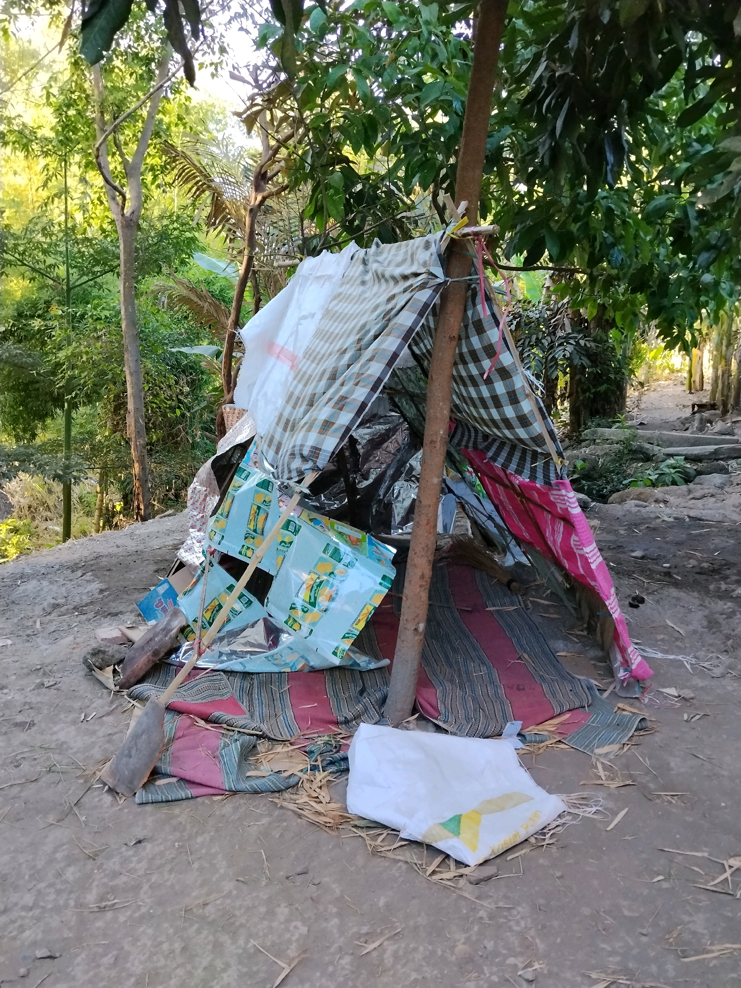 This is a medium shot, taken outdoors, of a makeshift shelter constructed from various fabrics and possibly tarps, supported by wooden poles. The shelter is situated on the ground, which appears to be dirt and sand, with scattered dry leaves and twigs. In the background, there is dense foliage and trees, suggesting a rural or semi-rural environment. A pathway with some steps can be seen leading away from the shelter into the greenery. The shelter itself is colorful, with striped fabrics in red, blue, and white, as well as patterned fabrics in green and white, and a light blue tarp with yellow graphics. There is also a white plastic bag in the foreground. No people are visible in the image. The scene is likely located in the city of Kejawan, Indonesia, based on the provided context.