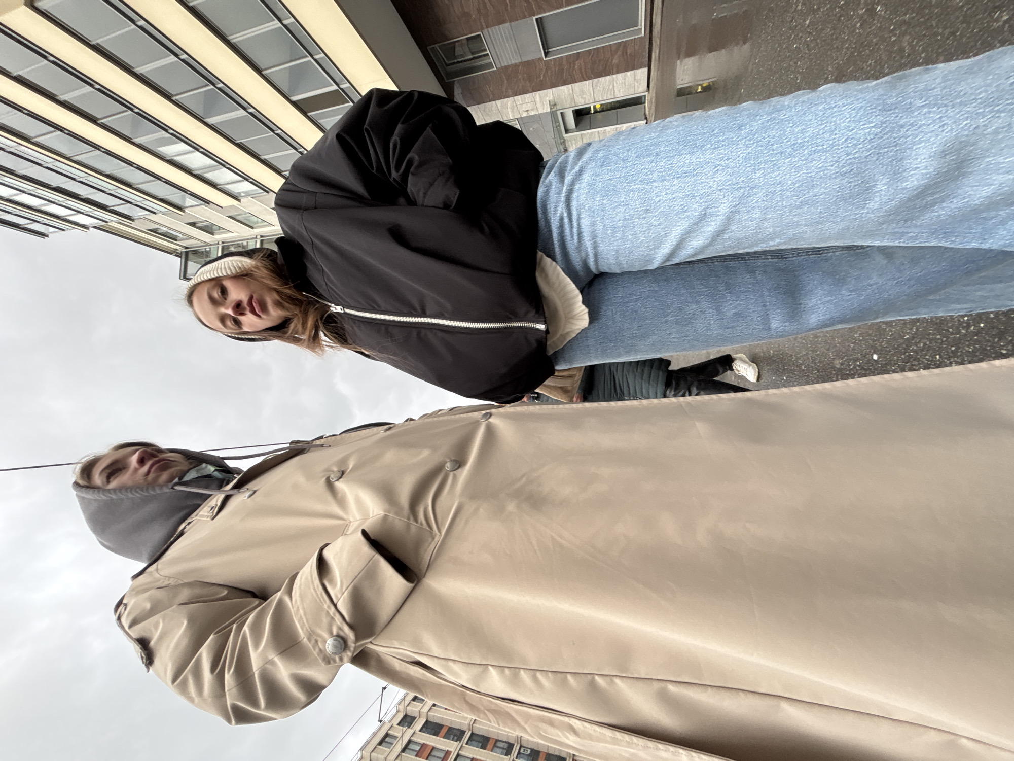 Two individuals are photographed in an urban setting of Unknown, Russia, from a low angle looking upwards. One person wears a black jacket and light blue jeans, topped with a white beanie. The second person is dressed in a tan trench coat with a gray hood. Buildings line the background, and the sky is overcast.