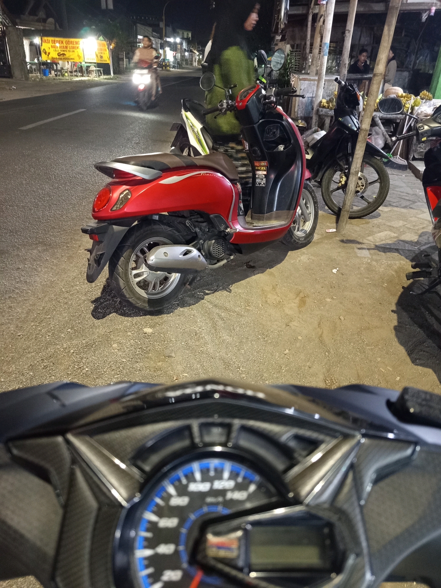 The image depicts a nighttime street scene in Wirowongso, Indonesia, characterized by an outdoor food stall on the left and motorcycles parked on the right. A bright yellow sign with text in Indonesian, advertising "NASI BEBEK GORENG NASI AYAM GORENG LELE TYME" (Fried duck rice, Fried chicken rice, Catfish time), is illuminated by fluorescent lights. Several tables and chairs are visible under the stall.
In the foreground, a red and black scooter is parked, its rear visible. Behind it, a black motorcycle is parked. Further down the street, another motorcycle with a rider is illuminated by its headlight, moving away from the camera. A person wearing a green hooded jacket is standing near the red and black scooter, partially obscuring the view of a black motorcycle parked next to it. Another person is visible in the background, behind a stall with bananas and other produce. The ground is a mix of asphalt and gravel. The image appears to be taken from the perspective of someone sitting on a motorcycle, with a portion of its dashboard visible in the bottom of the frame.