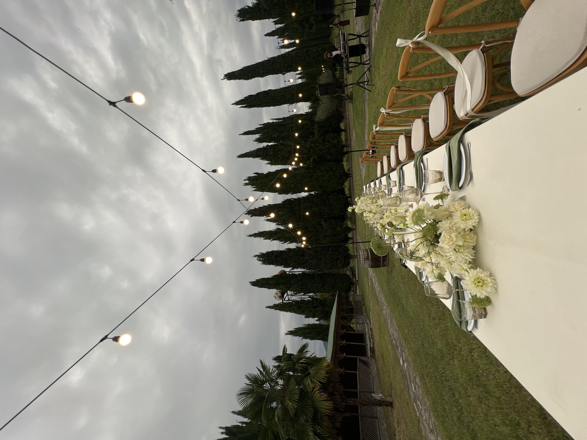 The image depicts an outdoor long table setting, likely for an event, in Napareuli, Georgia. The table is covered with a white tablecloth and adorned with white floral arrangements, place settings with glassware, plates, and green napkins. Wooden chairs with light-colored cushions are positioned along one side of the table. String lights with visible bulbs are suspended overhead, creating an atmospheric ambiance against a cloudy sky.
In the background, a row of tall cypress trees lines the landscape, with a rustic wooden structure and a palm tree to the left. Further in the distance, a person is visible standing by DJ equipment, suggesting music is being provided. The scene is set on a grassy area with a paved pathway winding through it. The overall environment appears to be a garden or estate, consistent with a rural Georgian setting.