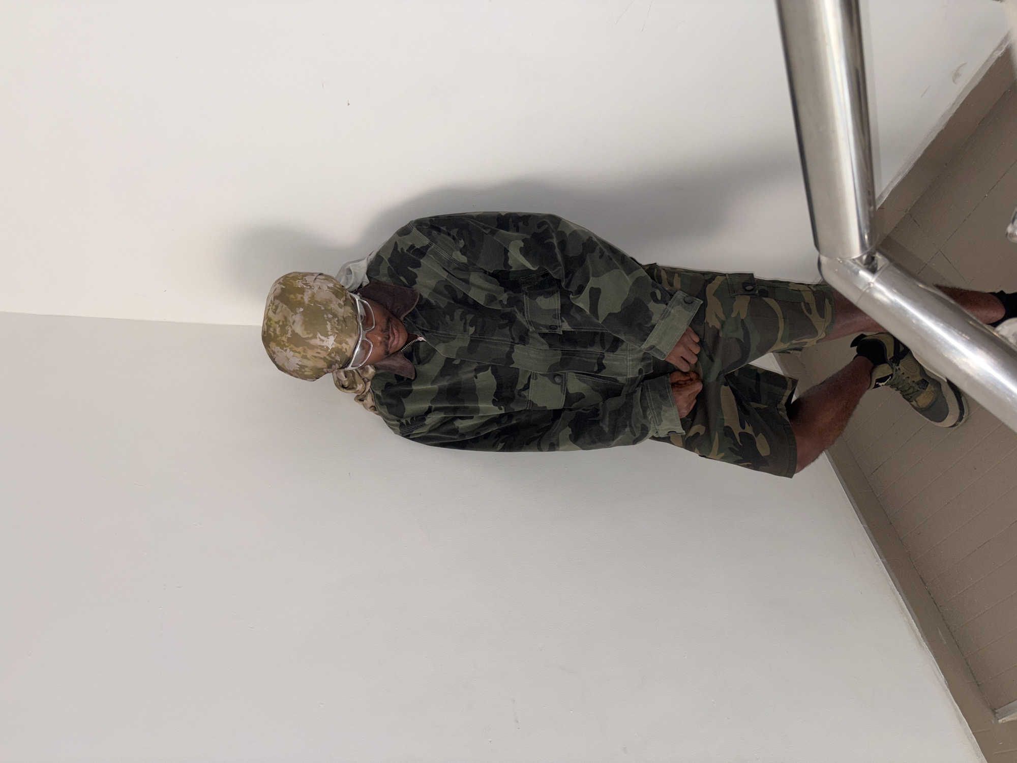 A man wearing camouflage clothing (jacket, shorts, and hat) and glasses is standing next to a silver metal handrail. The location is Igboefon, Nigeria. He is wearing athletic shoes. He has dark skin. The walls are white.