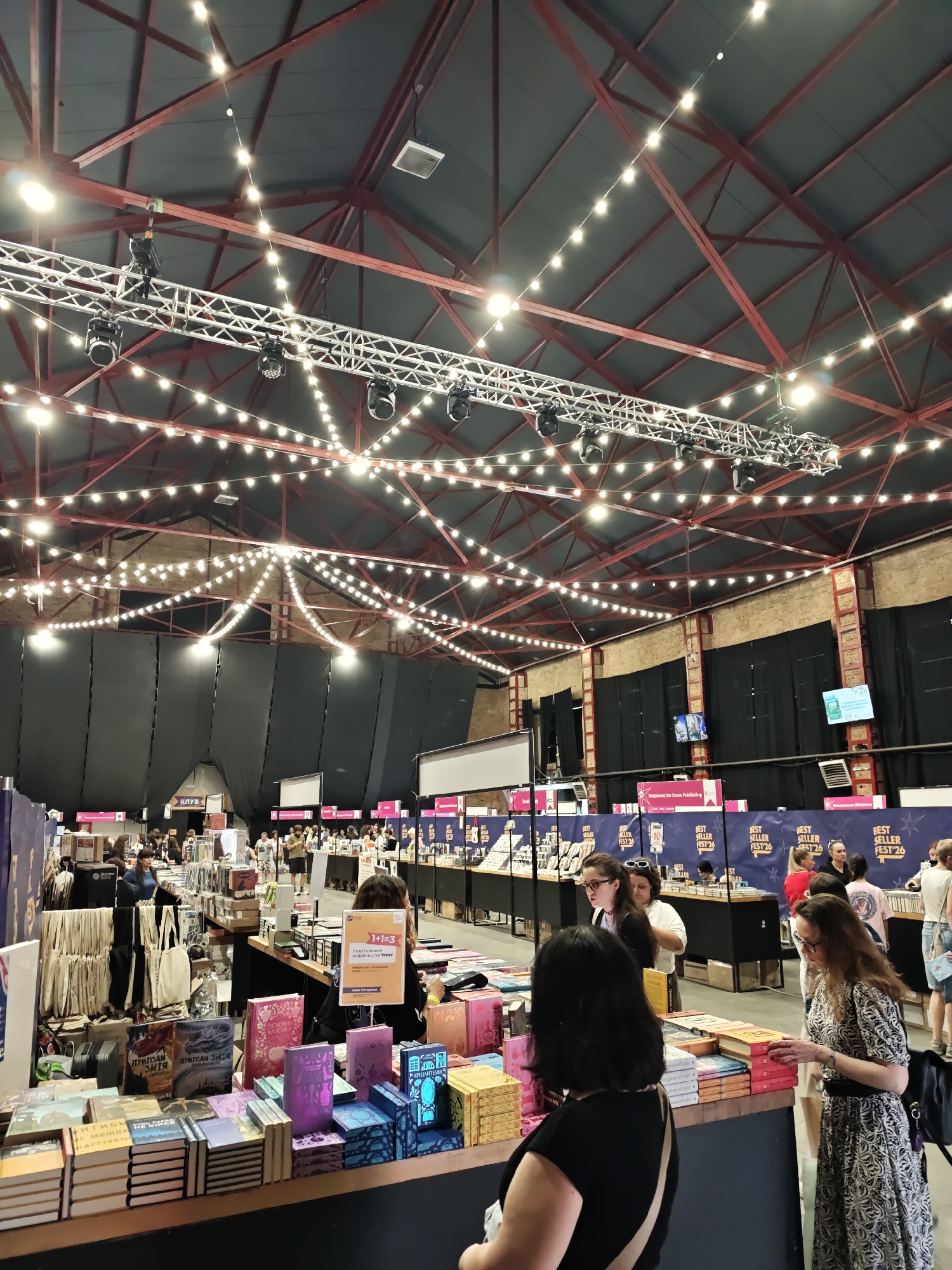 The image depicts an indoor book fair held at the Potocki Palace in Lviv, Ukraine. The scene is bustling with activity, featuring numerous people browsing and interacting at various stalls. The venue is characterized by its high, exposed ceiling with red beams and a network of decorative string lights, creating a festive atmosphere. Stalls are set up with tables laden with books, displayed in stacks and rows, catering to a wide range of interests. Several banners and signs are visible, some promoting "Best Seller Fest '26", indicating a literary event. People of diverse ages and genders are seen engaged in activities such as examining books, conversing with vendors, and walking through the aisles. The lighting is a mix of ambient overhead lights and the warm glow of the string lights. The overall impression is one of a vibrant and engaging cultural event.