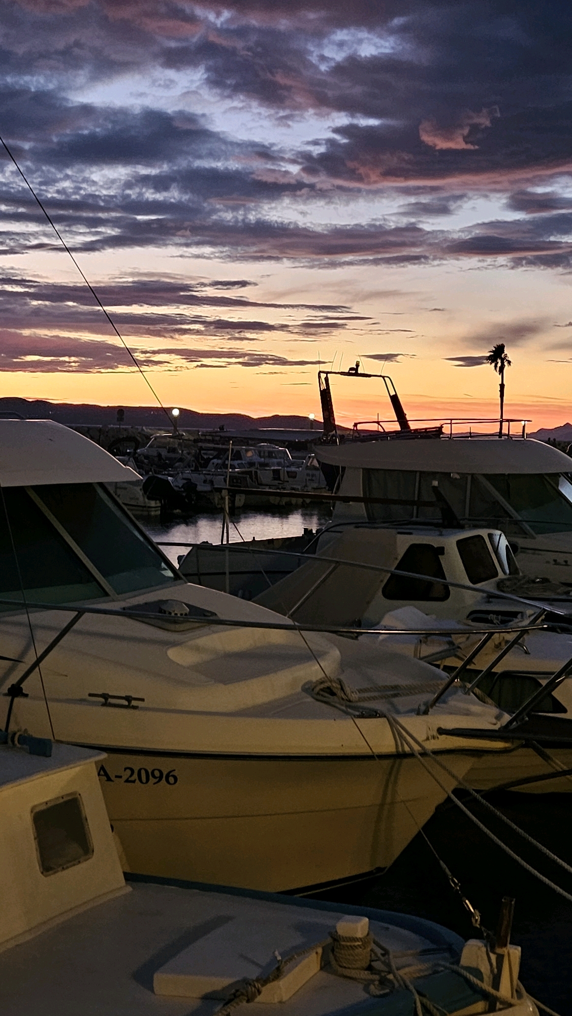 At sunset, several boats are docked in a harbor in Općina Tučepi, Croatia. The sky is filled with dramatic clouds in shades of purple, pink, and orange, illuminated by the setting sun. In the foreground, a white boat with the registration "A-2096" is prominently displayed. Other boats are visible in the background, some appearing to be larger vessels. Hills and a lone palm tree can be seen on the horizon. The water is calm, reflecting the colors of the sky. The scene depicts a peaceful evening in a coastal town.