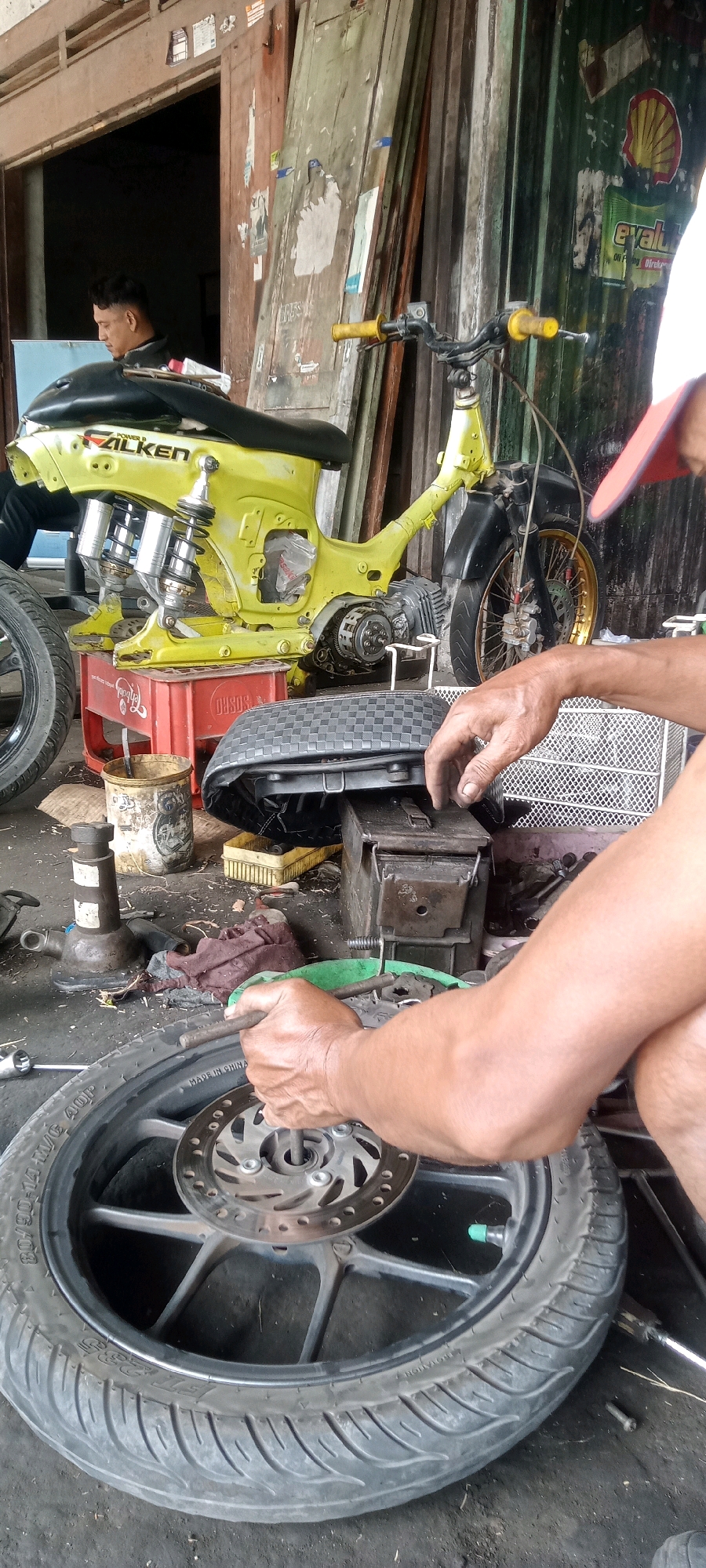 In Keteguhan, Indonesia, a mechanic is seen working on a yellow motorcycle frame, stripped down to its essential components, in what appears to be an outdoor workshop. The motorcycle, a Honda Cub or similar model, is supported by a red plastic crate. The engine is exposed, and the rear suspension, featuring dual shock absorbers, is prominent. In the foreground, the mechanic's hands are actively engaged in manipulating a motorcycle wheel, which has a visible brake disc. The wheel itself is a tire with a "MADE IN CHINA" inscription, suggesting its origin. Other objects in the scene include tools, a tire, a red crate, and a worn-out motorcycle seat resting on top of a metal box. The background reveals a rustic structure with wooden doors, some bearing faded posters and stickers, indicating a potentially long-standing establishment. The ground is littered with debris, typical of a workshop environment.