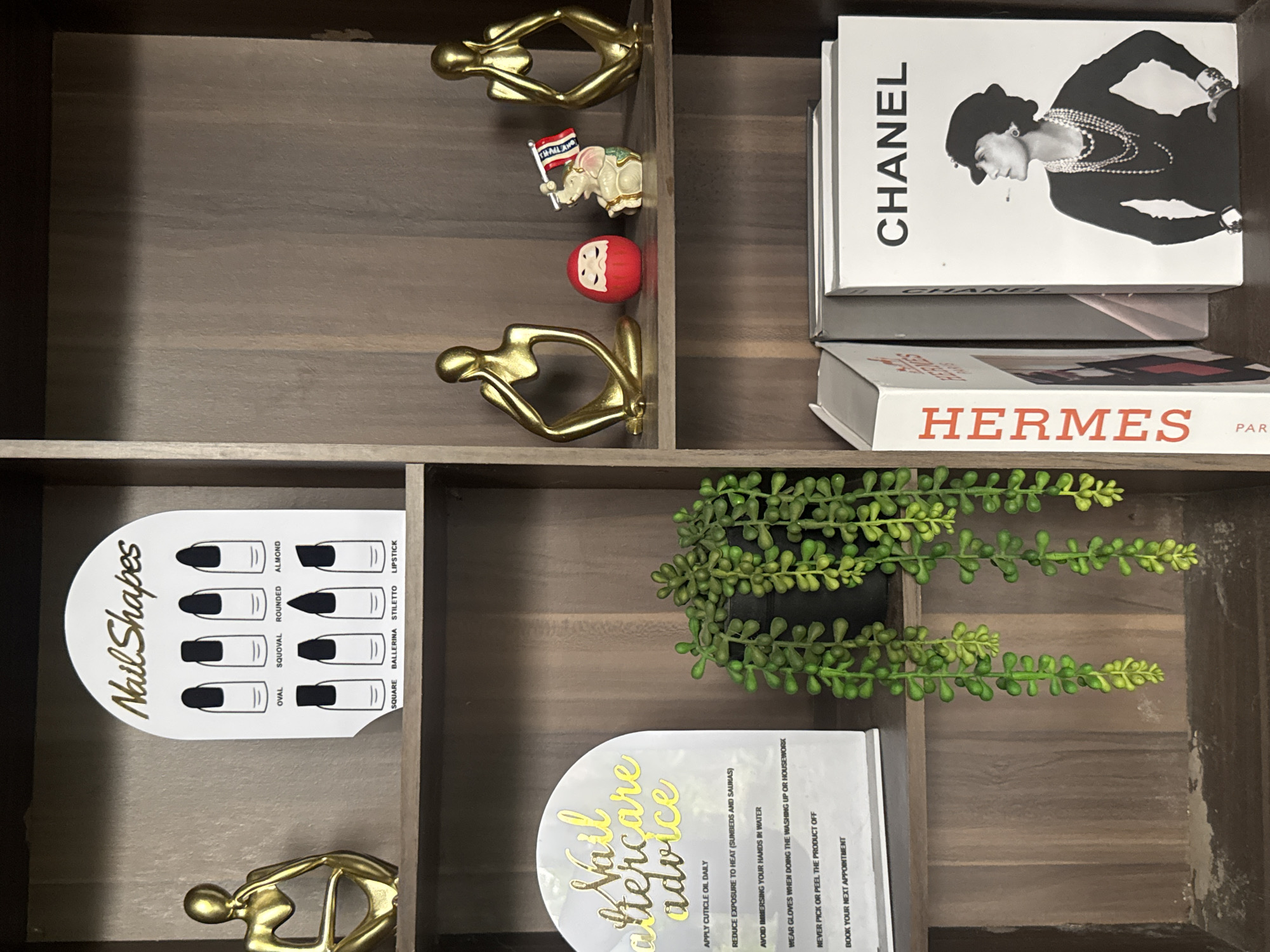 The image displays a dark brown, multi-tiered wooden shelving unit, likely situated in a professional setting such as a nail salon or beauty studio. The image itself provides no identifying information regarding its geographical location, including any city in the Philippines.
The top shelf, from left to right, features a small, abstract gold metallic sculpture of a seated figure with its hand to its chin. Next to it is an arched white sign titled "Nail Shapes" in gold script, illustrating eight distinct nail forms: Oval, Squoval, Rounded, Almond, Square, Ballerina, Stiletto, and Lipstick, each with a black stylized nail.
The middle shelf, from left to right, showcases a matching arched white sign titled "Nail aftercare advice" in gold script. Below the title, six bullet points of advice are visible: "APPLY CUTICLE OIL DAILY," "REDUCE EXPOSURE TO HEAT (SUNBEDS AND SAUNAS)," "AVOID IMMERSING YOUR HANDS IN WATER," "WEAR GLOVES WHEN DOING THE WASHING UP OR HOUSEWORK," "NEVER PICK OR PEEL THE PRODUCT OFF," and "BOOK YOUR NEXT APPOINTMENT." A cascading artificial plant with small, ovate green leaves in a black pot is positioned to the right of this sign. Further right on the same shelf are three decorative items: another abstract gold metallic sculpture of a seated figure with its hand to its face, a small red Daruma doll with a white face, and a small ceramic elephant holding a miniature flag that reads "THAILAND." On the far right, a third abstract gold metallic sculpture depicts a seated figure with hands covering its ears.
The bottom shelf, from left to right, displays two vertically oriented large format books. The left book has "HERMES" printed in red text on its spine. The right book, white, features "CHANEL" in black capital letters at the top, above a black and white portrait photograph of Coco Chanel, depicted in profile wearing pearls, a hat, and holding what appears to be a cigarette. The floor of this bottom shelf shows some light dust accumulation.