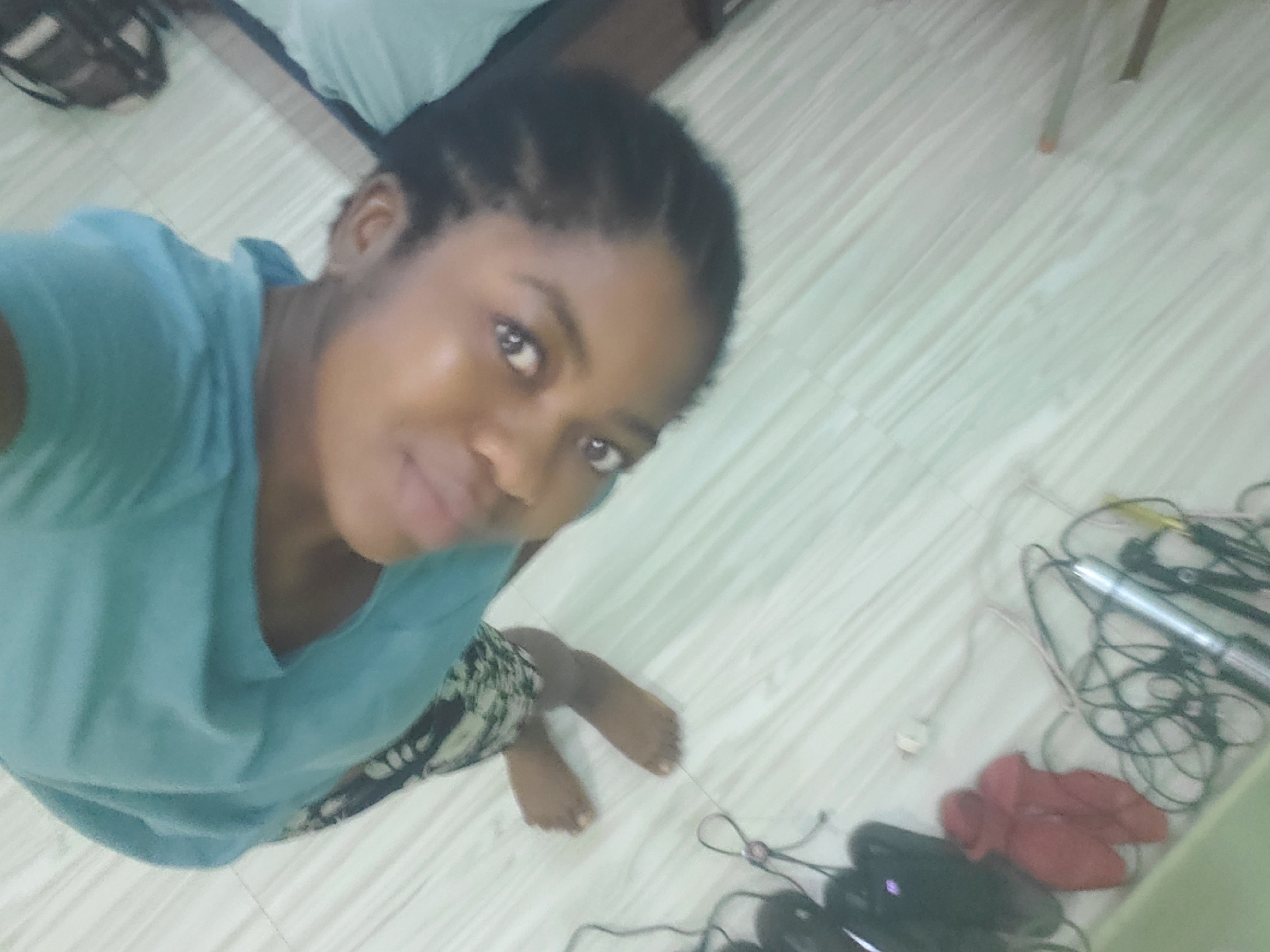 A high-angle shot shows a young Black woman lying on her back on a floor with a wood-like pattern. She is wearing a blue t-shirt and a patterned skirt. Her head is tilted to the side, and she is looking towards the camera. Her hair is braided. Her feet are visible, and she is resting on what appears to be a light-colored surface. In the background, there are several electronic devices and wires. The scene appears to be indoors, possibly in a bedroom or living area. There is no indication of the specific city of Barden Yana, Nigeria.