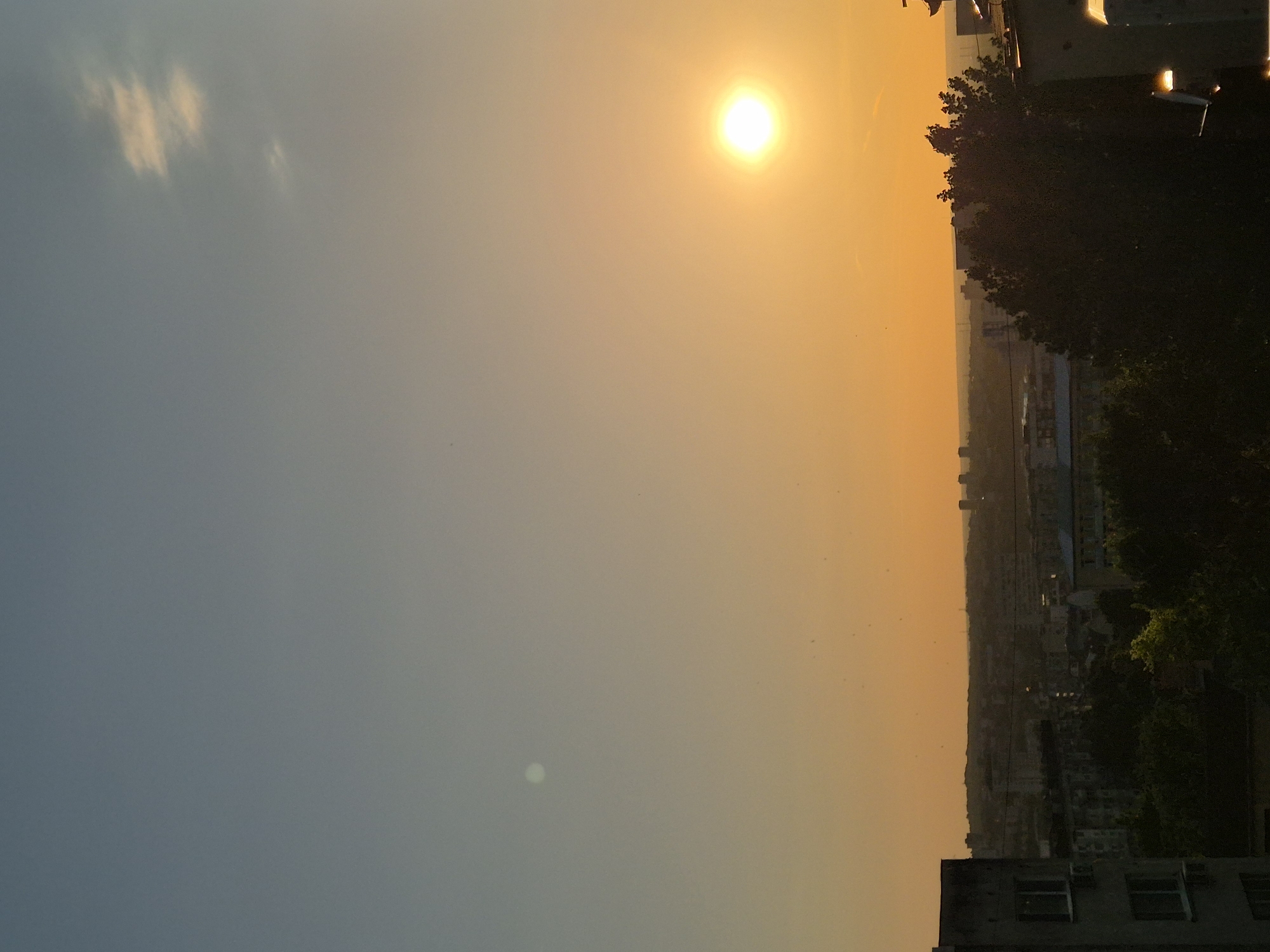 The image shows a sunset view of a city. The sun is high in the sky, casting a warm, orange glow over the city. The sky transitions from blue to orange as it gets closer to the horizon. There are trees and buildings silhouetted against the orange sky. The location is the city of Unknown, Unknown.