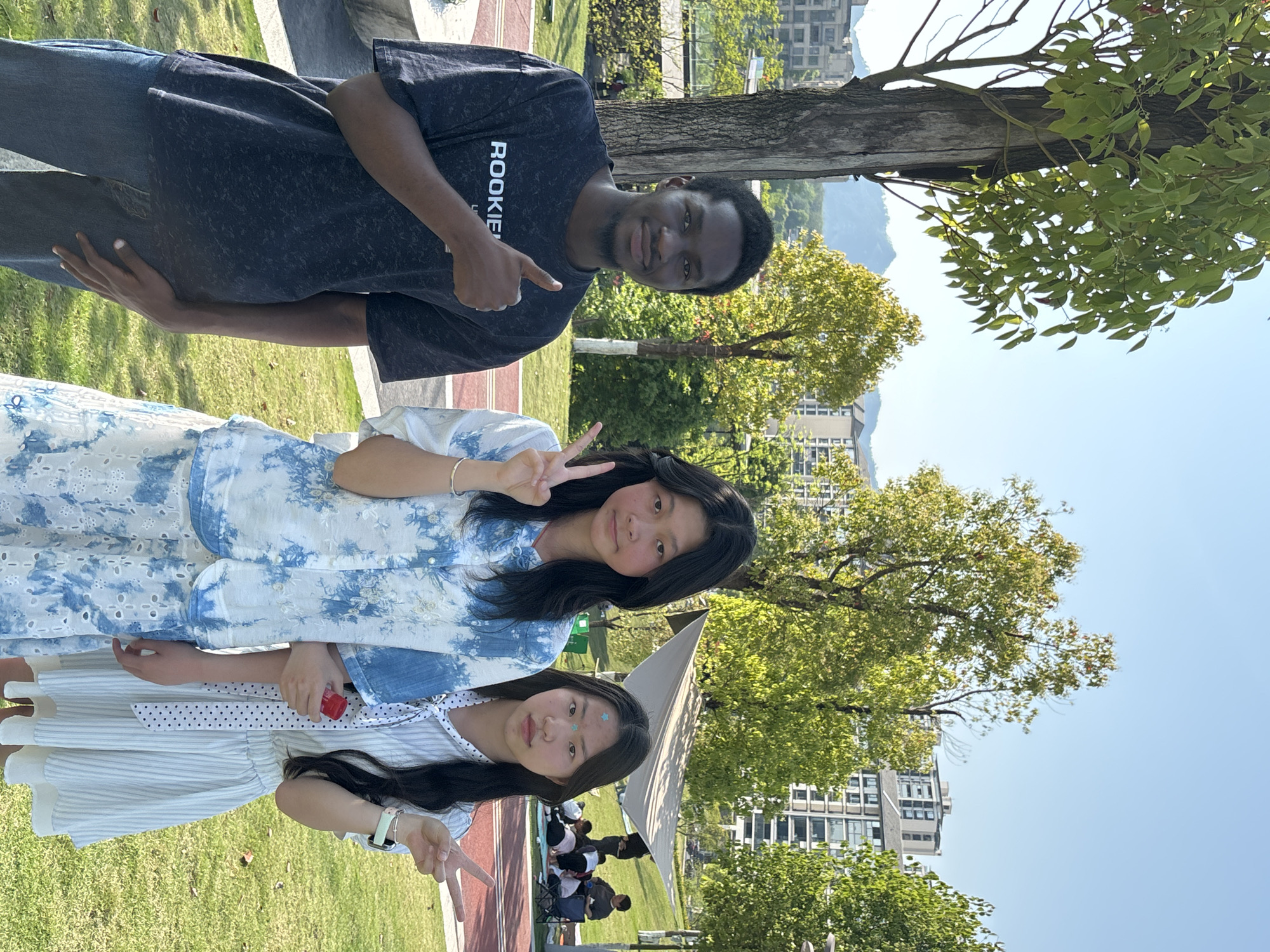A medium shot depicts three individuals, two young Asian females and one Black male, against a backdrop of an urban park setting. The two females are positioned at the bottom-center of the image, while the male stands above them.
The female on the bottom is wearing a white dress with blue stripes, and a small star-shaped design on her forehead. The female above her has on a blue and white patterned top. Both are making the "peace" sign with their hands. The male wears a dark-colored shirt that appears to have the word "ROOKIE" printed on it. He is pointing to the two girls.
The setting includes grass, trees, buildings, and what appears to be a small tent. People are gathered and sitting on the ground near the tent, suggesting a public or recreational space. Given the presence of distinct urban architectural features, trees, and a clear sky, this image likely originates from a public park in Jiangjin, China, as specified.
