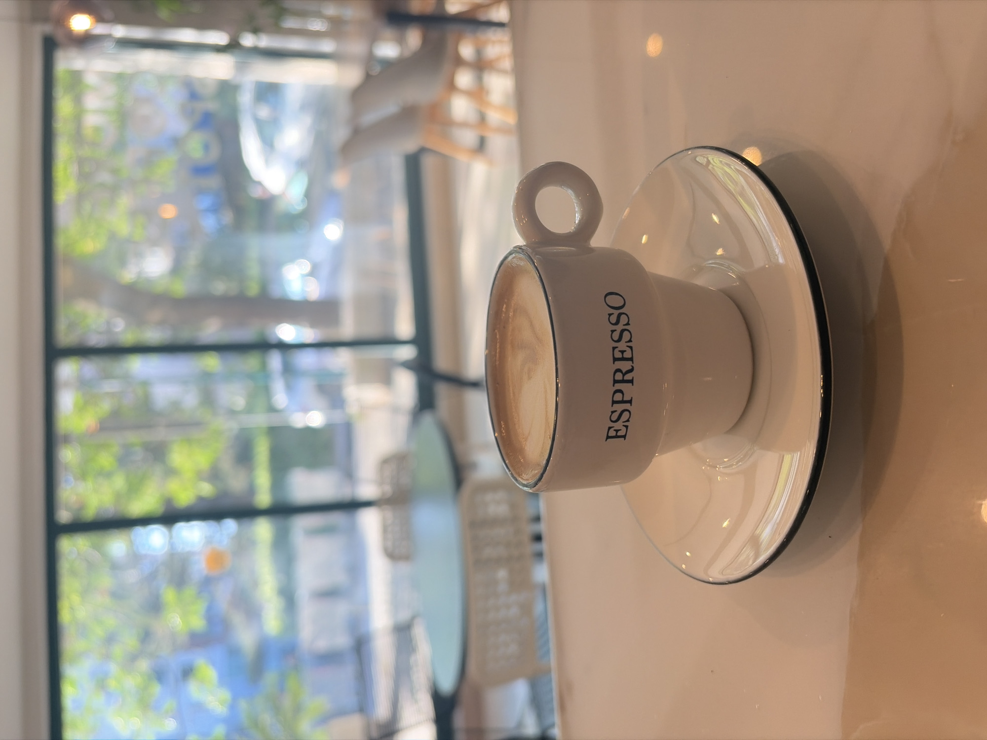 A close-up, eye-level shot features a white espresso cup and saucer on a light-colored, reflective surface. The cup has the word "ESPRESSO" printed in black lettering. The background, visible through a window, is blurred and shows greenery, trees, and buildings, suggesting an outdoor cafe or restaurant setting in Budva, Montenegro. The overall ambiance is bright and airy, with soft lighting.