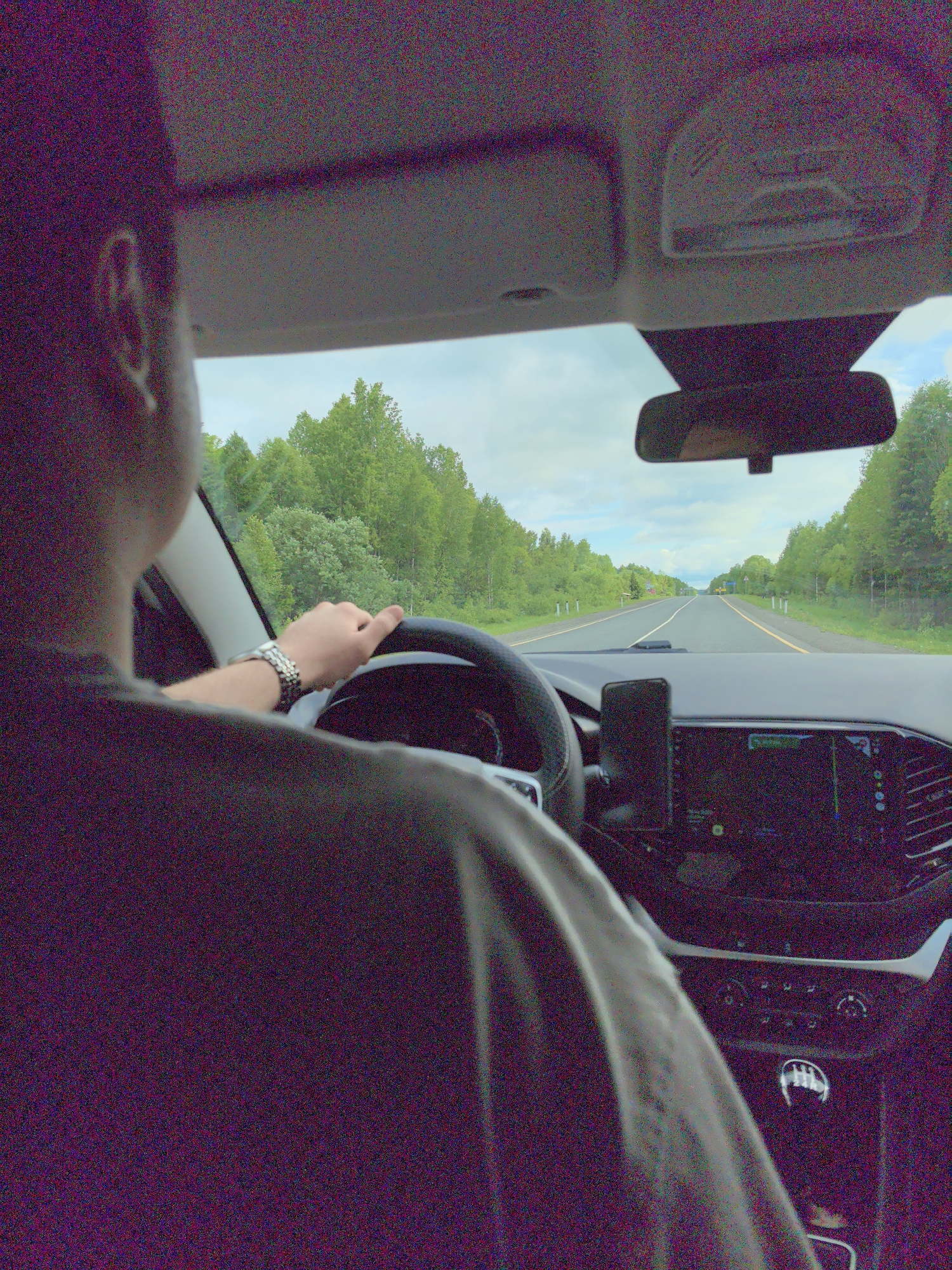 The media file captures an interior view from a car, looking forward through the windshield. A person, seen from behind, occupies the driver's seat. Their back of the head and right arm are visible, with the hand grasping the steering wheel. A silver-colored watch is worn on their left wrist.

The car's interior features a modern dashboard. A large infotainment screen, appearing to display a navigation system with faint icons, is visible in the center console. A smartphone is mounted to the right of the steering wheel. The rearview mirror is positioned centrally at the top of the windshield. Above, the overhead console with light controls and a passenger-side handle on the A-pillar are partially visible. A symbol resembling an "R" (for Reverse) is faintly discernible on the gear selector knob.

Outside the car, a two-lane asphalt road extends into the distance under a bright, overcast sky with patches of blue. The road is clearly marked with white dashed lines in the center and solid yellow lines along the shoulders. Dense green trees and foliage line both sides of the highway, creating a continuous forested landscape. Several other vehicles can be seen in the distance on the road ahead.

The image depicts a daytime drive through a rural or semi-rural area, specifically in Anninsk, Russia. The photograph exhibits significant digital noise or grain throughout.