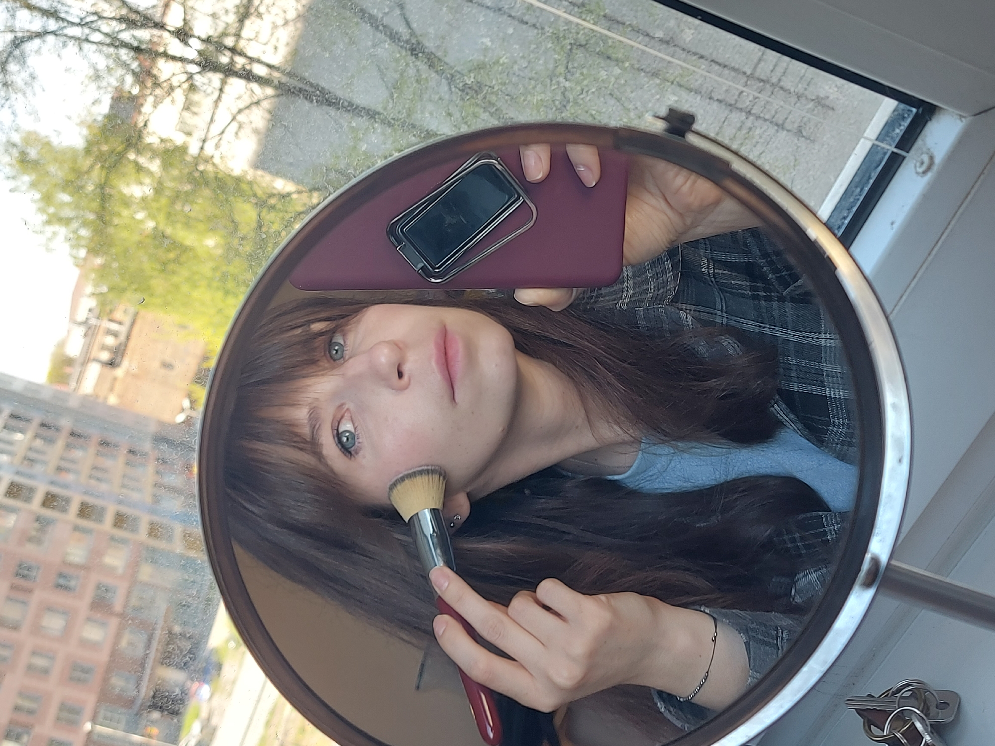 A young woman is shown in a mirror selfie. She is holding a makeup brush to her cheek. In the background reflected in the mirror, buildings are visible, suggesting an urban environment. The presence of what appears to be a window frame indicates she is inside a building. Based on the architectural style of the buildings in the reflection, the location may be Saint Petersburg, Russia, characterized by its European urban landscape. The woman holds a smartphone against the mirror surface. Keys lay close to the bottom right of the mirror base. She is wearing a blue top under a gray and black checkered button down. A silver bracelet decorates her wrist.