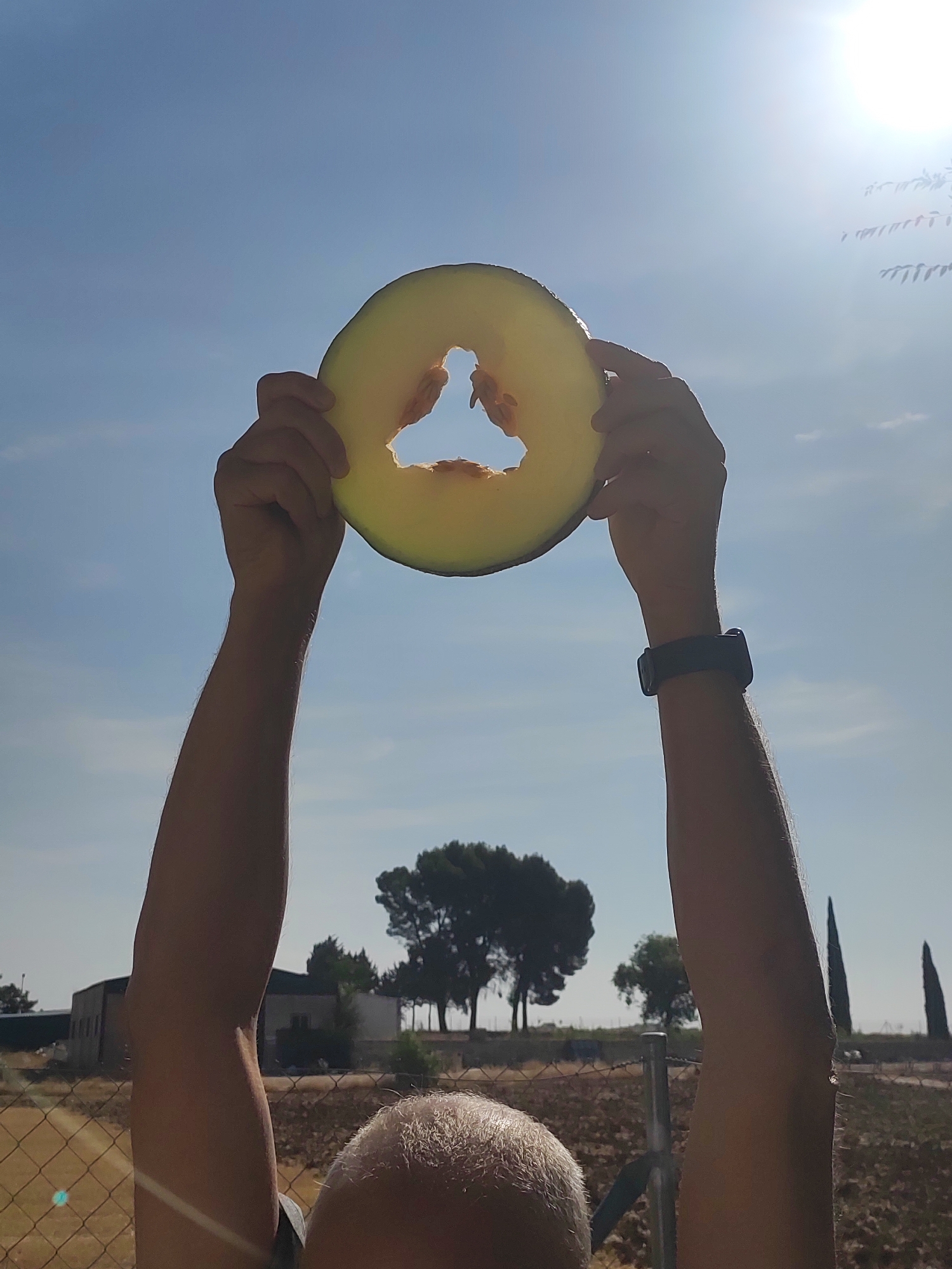 The image shows a person holding a slice of melon against a bright blue sky. The melon slice is roughly circular with a triangular hole in the center. The person's arms are raised, and their hands are visible holding the melon. The background shows a rural landscape with trees, a building, and a fence. The sun is visible in the upper right corner, creating a lens flare effect. The location is stated to be Villarejo De Fuentes, Spain.