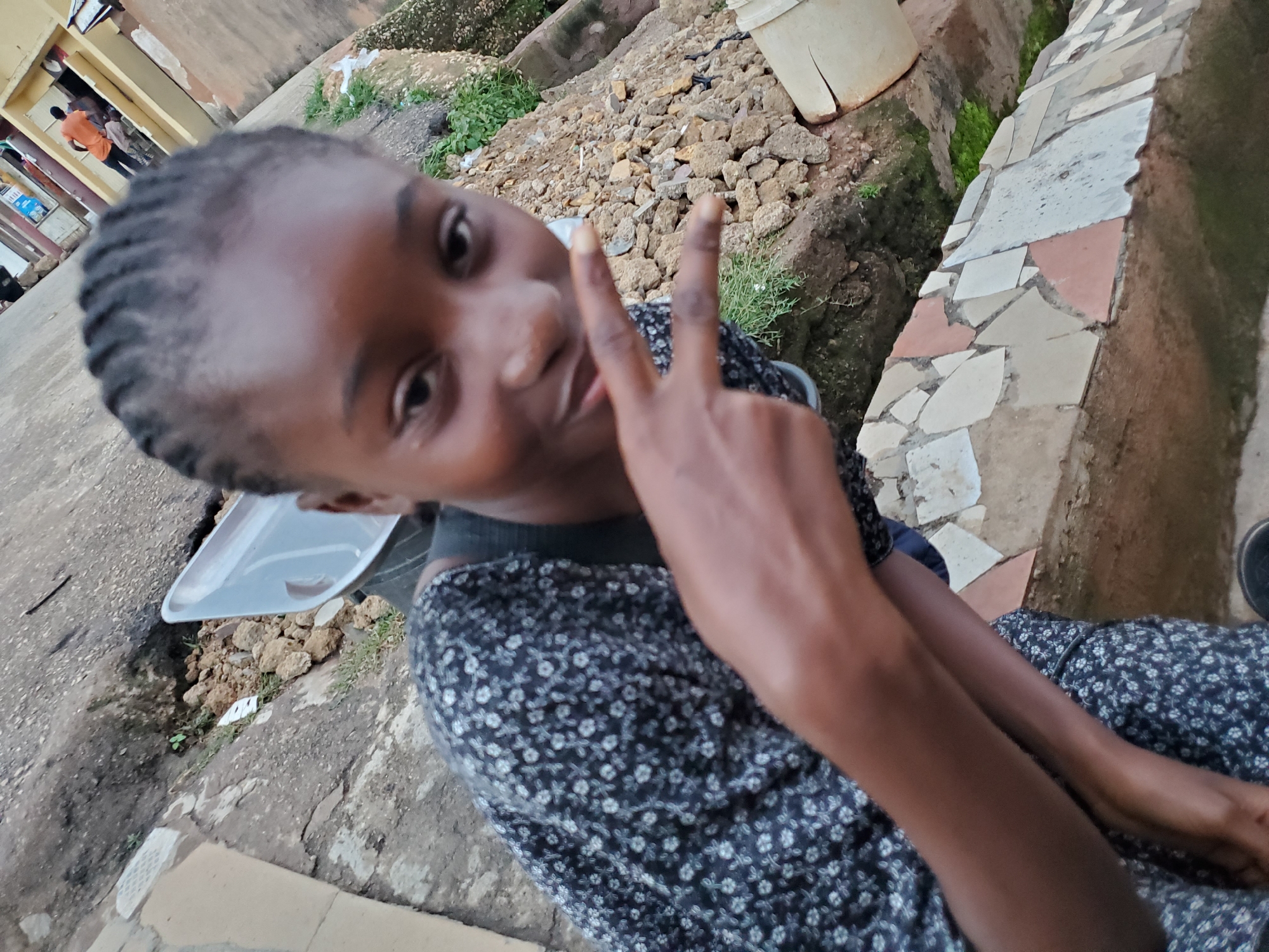In this close-up, slightly angled shot taken in Jos, Nigeria, a young person with braided hair smiles at the camera, making a peace sign with their right hand. They are wearing a dark blue dress with a white floral pattern. The background suggests an outdoor, somewhat unkempt environment. To the left, a pale yellow building with an open doorway is visible, with at least two figures in orange and dark clothing standing inside. In the mid-ground, a pile of rough stones and some greenery are present, next to a white bucket. Further to the right, a narrow trench is lined with mosaic tiles in shades of white, pink, and beige. The ground appears to be a mix of dirt and gravel. The overall impression is of a casual, everyday scene in a residential area. The lighting suggests it is daytime, with no specific indication of the weather beyond the general outdoor setting. The young person's expression is cheerful and engaging. No visible text is present in the image.