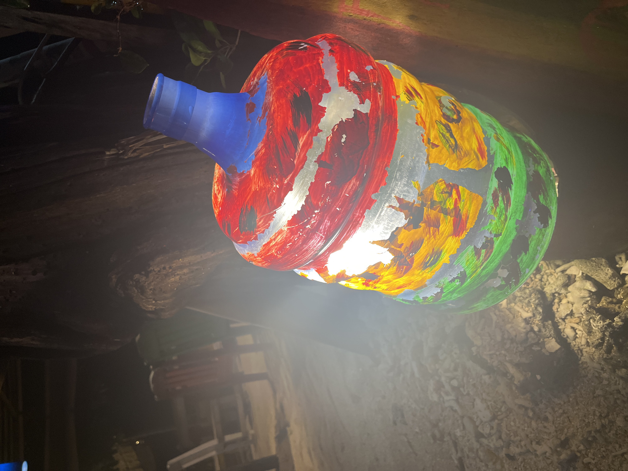 An illuminated, repurposed plastic water bottle, painted with red, yellow, and green hues and a blue neck, is positioned in an unknown indoor location. The background is largely obscured, showing only glimpses of what appears to be wooden supports and a concrete or rock surface beneath the bottle. The light source within the bottle shines brightly. The location is Unknown, Indonesia.