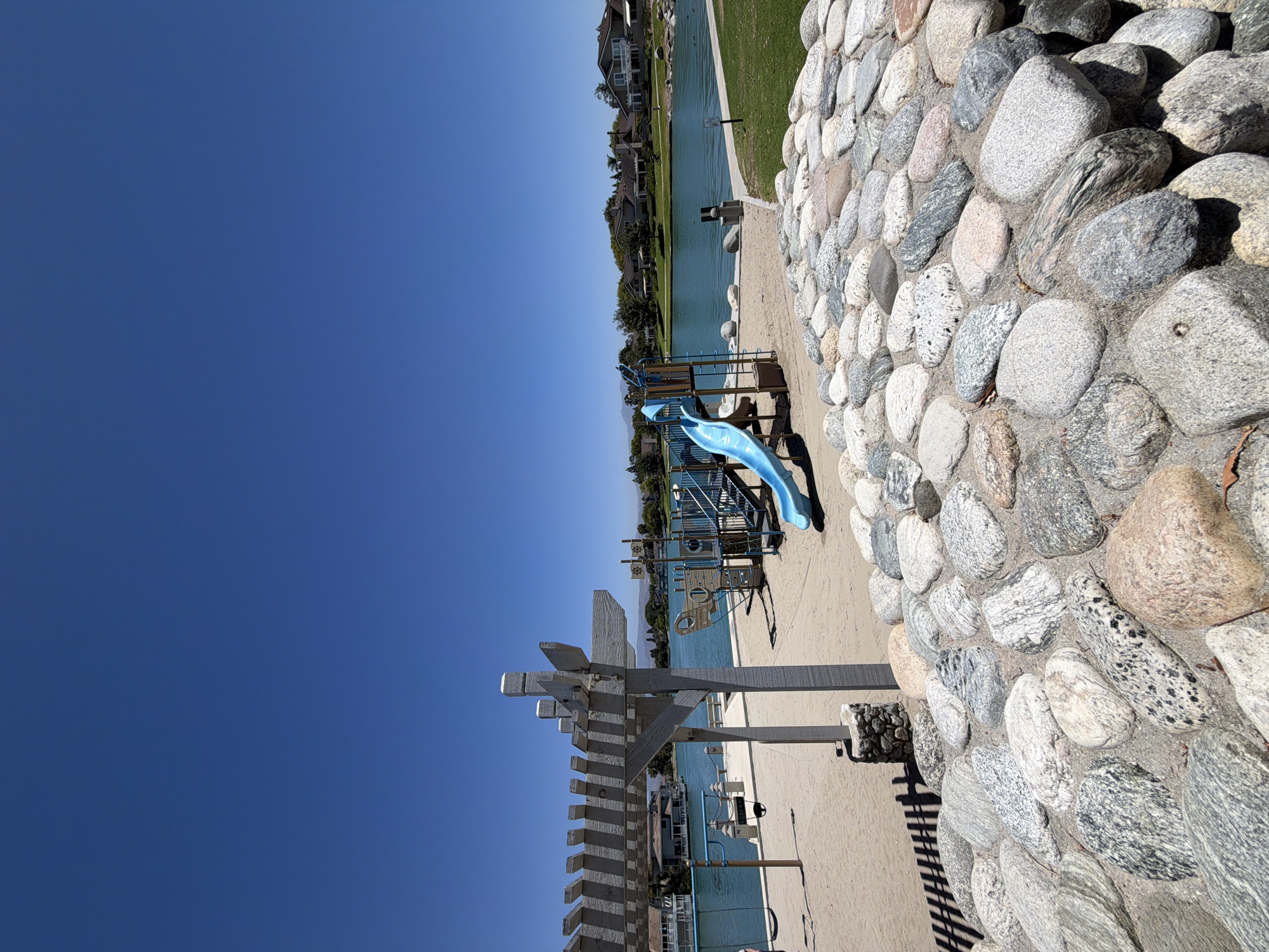 This image depicts a sunny day at a waterfront park in Irvine, California, USA. The foreground is dominated by a large arrangement of river rocks, varying in size and color, embedded in sand. Behind the rocks, a sandy playground area is visible, featuring a blue slide and a multi-level play structure with climbing elements, resembling a ship. Swings with dark seats are suspended from a wooden pergola-like structure.
The middle ground showcases a calm, blue body of water, likely a lake or lagoon, with houses visible on the opposite shore. These houses appear to be single-family residences with pitched roofs and various architectural styles. Trees and landscaping dot the shoreline.
The background is characterized by a clear, bright blue sky. The overall impression is one of a peaceful, recreational environment, likely a community park designed for outdoor activities and relaxation by the water.