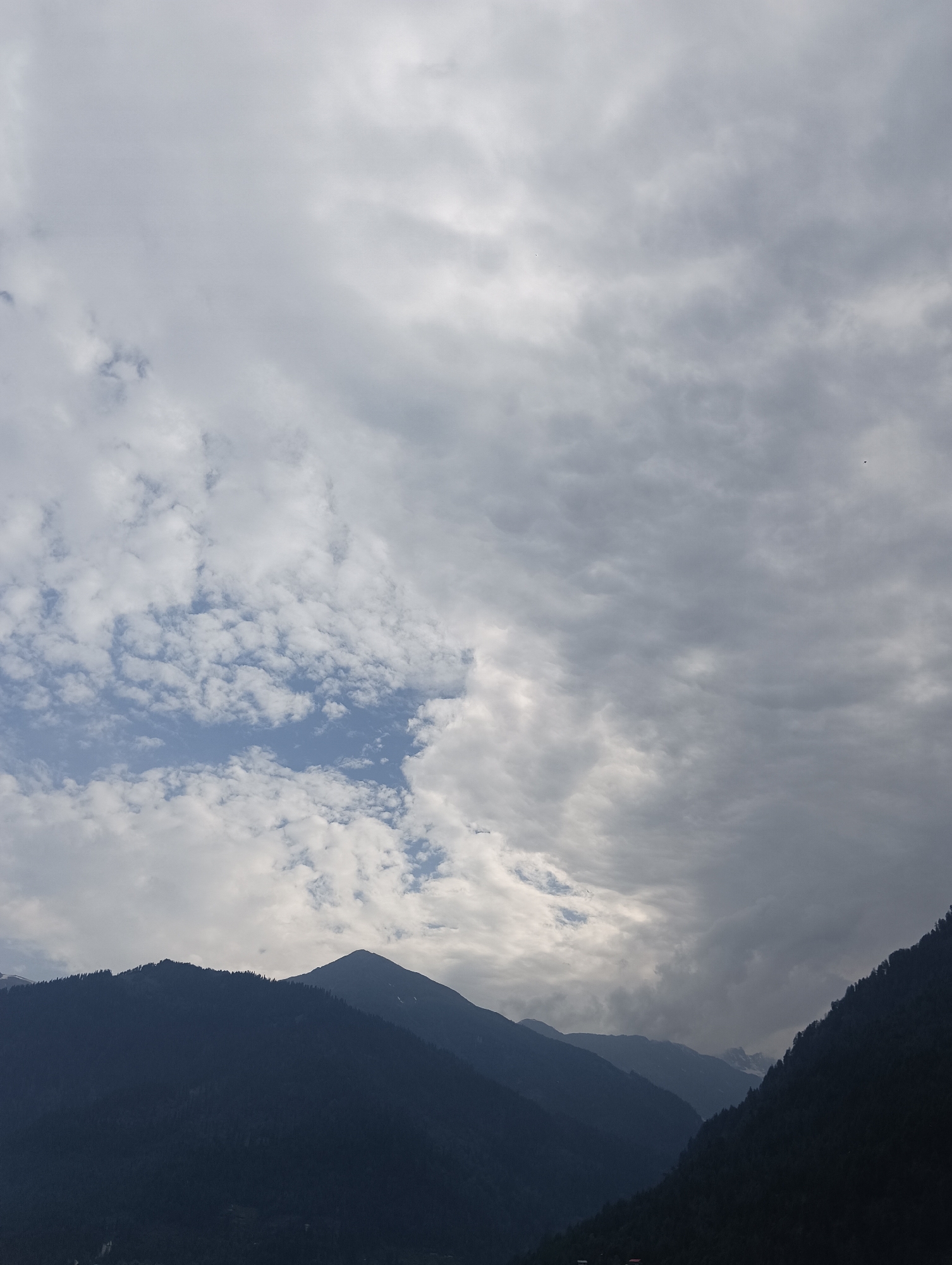 This media file depicts a majestic mountain landscape under a cloudy sky. The scene is dominated by the imposing silhouettes of dark, tree-covered mountains that fill the lower portion of the frame. The peaks are rugged and appear to stretch across the horizon. Above, the sky is a dynamic canvas of clouds, ranging from light, scattered white puffs to darker, more ominous gray masses that suggest impending or recent rain. Patches of blue sky peek through in some areas, indicating a break in the clouds. The overall impression is one of a grand, natural setting, possibly at a high altitude. The light suggests it could be late afternoon or early morning, with the sun's rays struggling to penetrate the cloud cover. There are no visible people or man-made objects, emphasizing the wildness of the location. The mood is serene yet powerful, evoking a sense of awe at the scale of nature. The location is contextualized as Vashisht, India, a place known for its mountainous terrain.