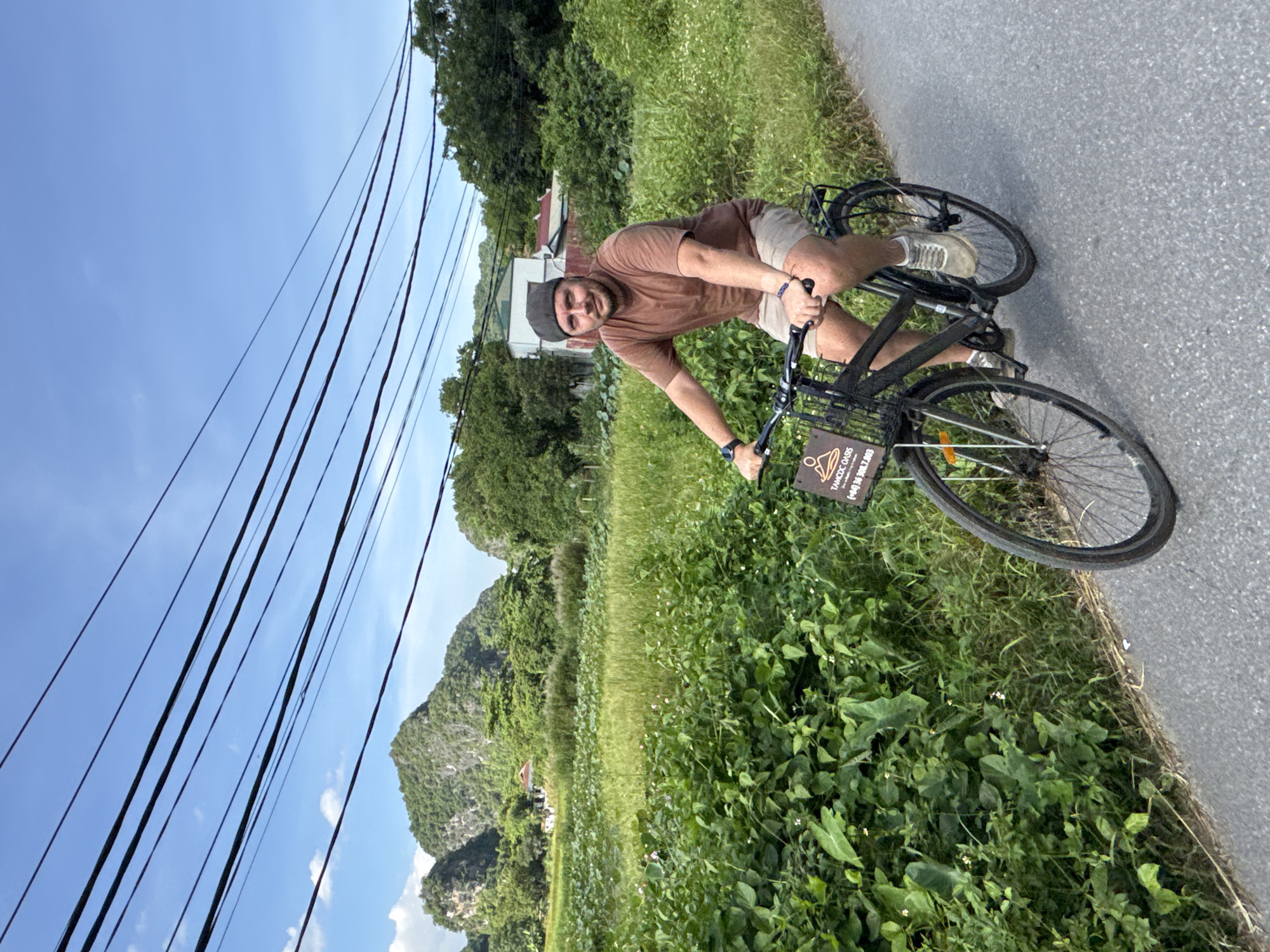 A man in a brown t-shirt and tan shorts rides a black bicycle on a paved road. He wears a grey flat cap and white sneakers. The bicycle has a basket on the front with a sign that reads "TAMCOC OASIS (+84) 36 308.7803". He is looking towards the camera. The road is bordered by lush green foliage on the left and grass and trees on the right. In the background, there are rolling green hills and a few houses. The sky is blue with scattered clouds and is crossed by several power lines.