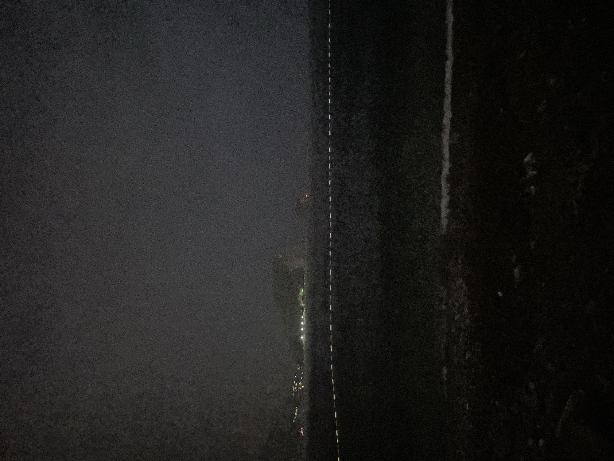 The image depicts a dark, slightly foggy night scene. The most prominent features are two distinct vertical lines. The left line is a hazy, almost translucent boundary separating a dark, undefined area on the far left from a more defined middle ground. Scattered lights, appearing as small, bright dots, are visible along this middle ground, suggesting human habitation or activity. One red light is particularly noticeable. To the right of these lights is a distinct, broken white line, likely a road marking, running vertically through the image. This line separates the middle ground from a much darker, denser area on the far right, which appears to be textured and perhaps natural, like a dense bush or trees. The overall impression is one of looking across a dimly lit landscape at night, possibly from an elevated position, with the fog or haze muting the details and creating an atmospheric, somewhat mysterious feel. The time of day is clearly night, and the low visibility suggests a foggy or misty condition. There is no visible text in the image.