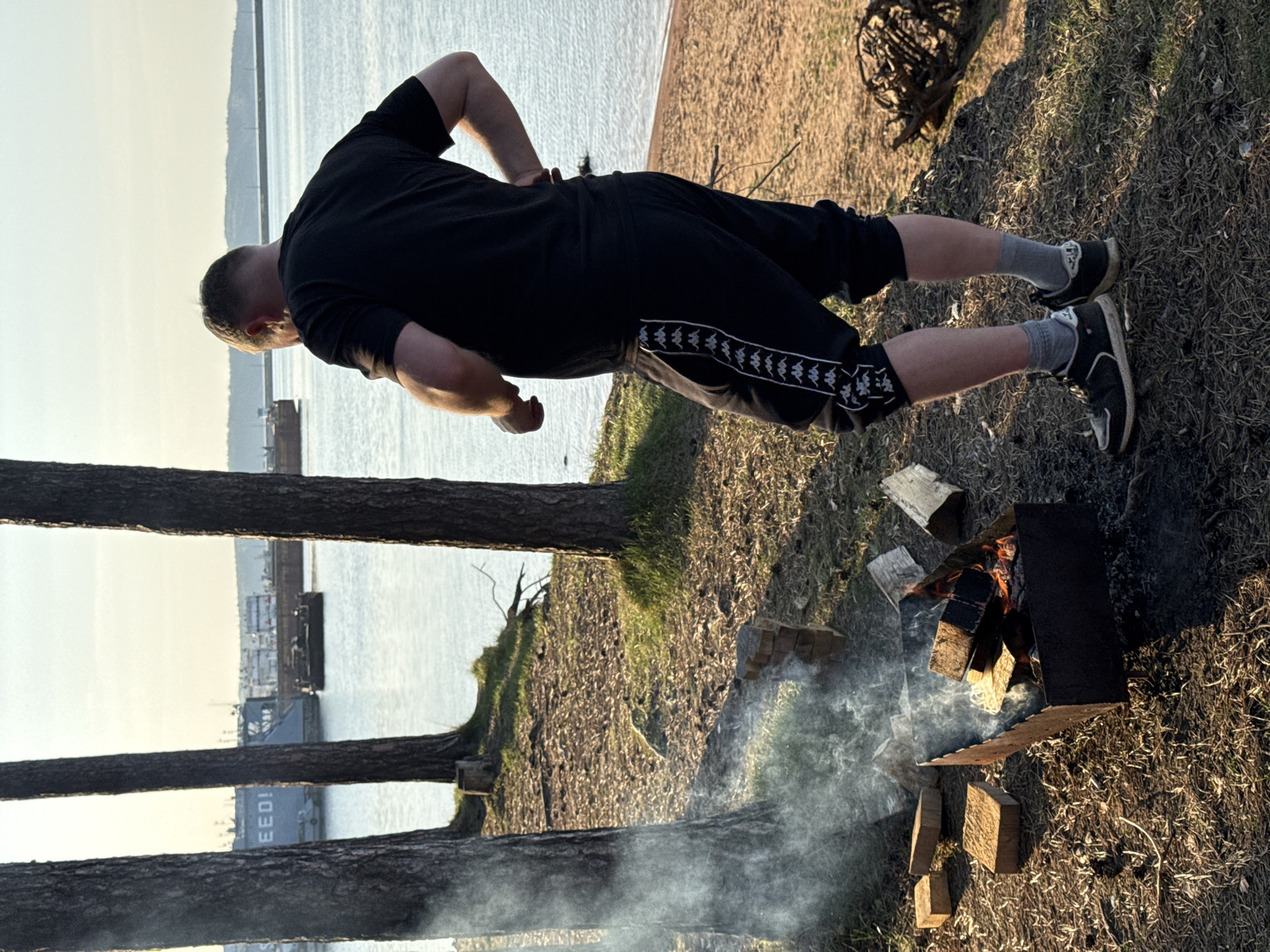 A person is standing next to a fire pit on a grassy area. The person is wearing a black shirt and black shorts with white stripes, gray socks, and black sneakers. The fire pit is made of wood and bricks and has smoke rising from it. In the background is a body of water with a dock and a ship. The location is Unknown, Unknown.