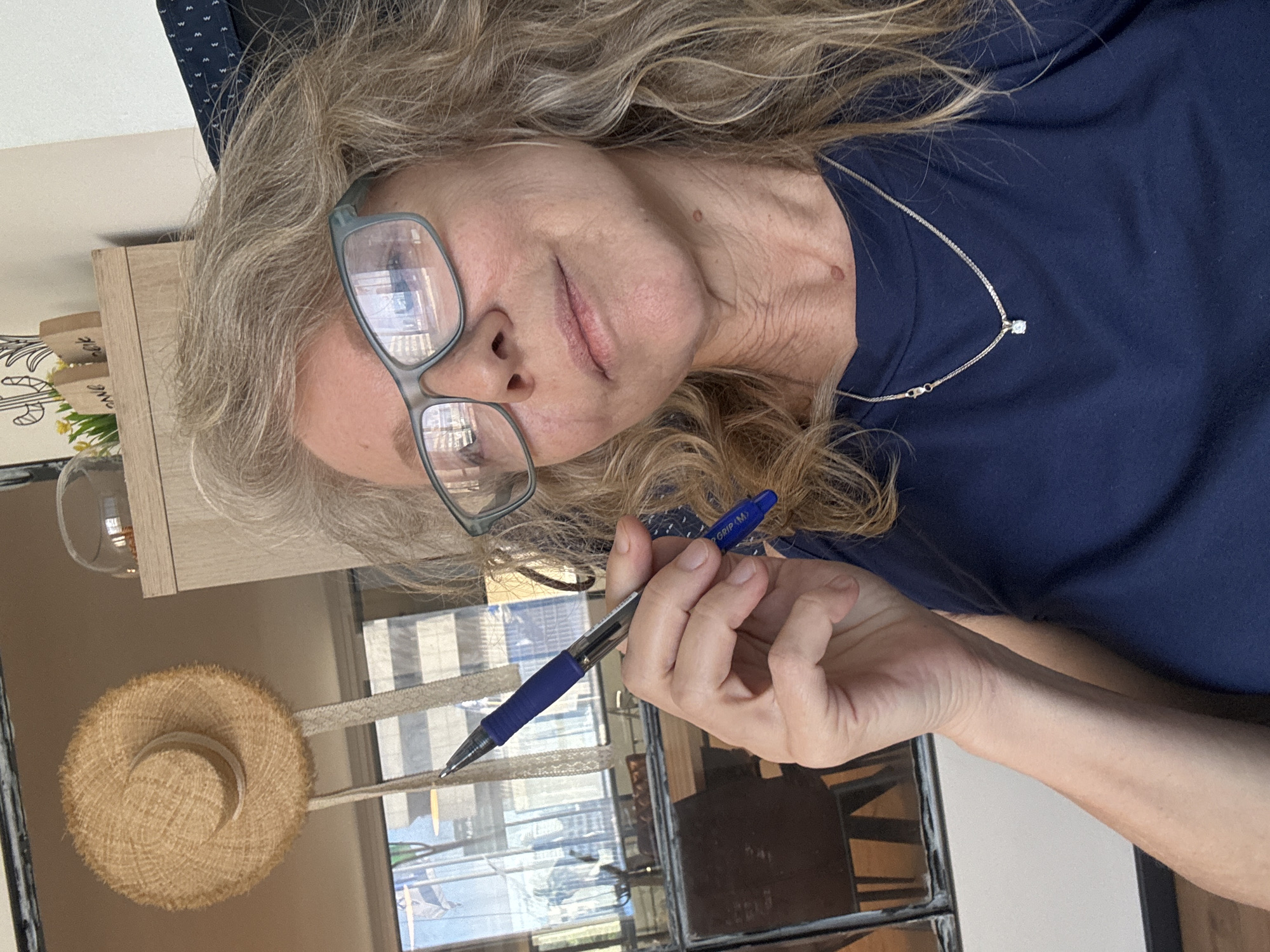 A woman with blonde hair, wearing glasses and a dark blue t-shirt with a silver necklace, is holding a blue pen. She is indoors, possibly in Tel Aviv, Israel, based on the reflection of buildings in the window. A straw hat is visible in the lower left corner, and there is a decorative item on a shelf behind her. The scene appears to be in a well-lit room.