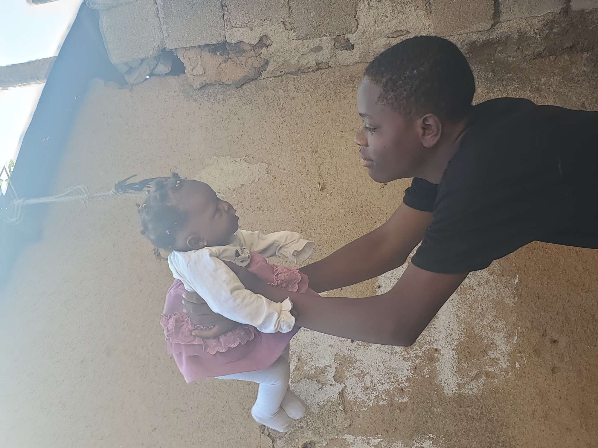 The image depicts an intimate moment between two individuals, an older child or young adult and a baby, in an outdoor setting in Jos, Nigeria. The older person, with dark skin and short dark hair, is wearing a black short-sleeved top and gently holding the baby. Their gaze is fixed on the baby with a tender expression. The baby, also with dark skin and small braids, is dressed in a white long-sleeved top and a pink ruffled dress over white leggings, and is looking up at the older individual, fostering a sense of connection.
The background features a rustic textured wall, likely constructed from concrete blocks or plastered earth, showing signs of wear and discoloration. The ground below is a similar earthy, textured surface. Bright natural light, entering from the upper left, indicates it is daytime, likely under clear, sunny weather conditions. A rope or string is visible in the upper left portion of the frame. There is no visible text in the image.