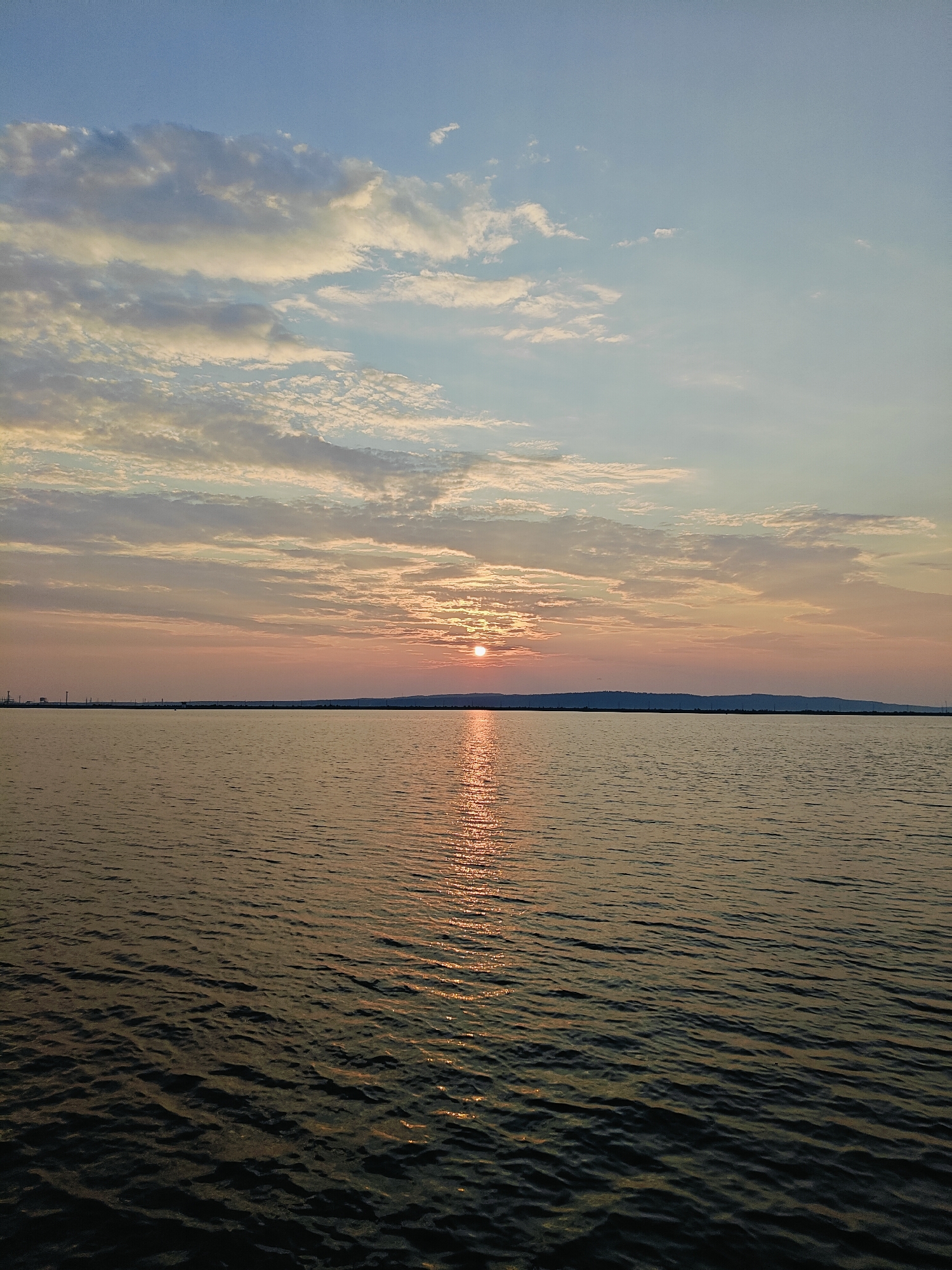 A wide shot captures a serene sunset over a body of water. The sun, a small, bright orb, is partially obscured by a strip of distant land, casting a shimmering golden reflection across the rippling surface of the water. The water itself is a deep, muted tone, with small waves creating patterns of light and shadow.
The sky is a canvas of soft colors. The upper portion is a clear, pale blue, transitioning to shades of pink, orange, and yellow closer to the horizon. Wispy clouds are scattered across the sky, catching the warm light of the setting sun and adding texture to the scene. Some of the clouds are tinged with white, while others have a darker, more dramatic hue.
Along the distant horizon, a dark, low-lying landmass is visible, punctuated by the silhouettes of what appear to be industrial structures or power lines. The overall impression is one of tranquility and natural beauty, with the sunset creating a peaceful and slightly melancholic mood. The time of day is clearly sunset. There is no visible text in the image. The location context of Chaikovsky, Russia, suggests this could be a view over the Kama River or one of its reservoirs.