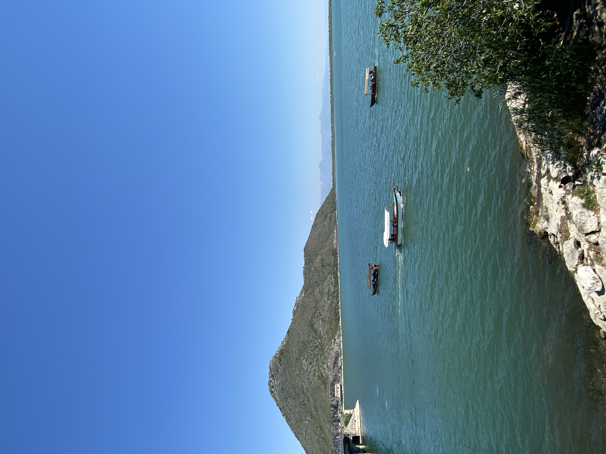The image depicts the landscape in Kruševica, Montenegro. A large body of water, possibly a lake or river, dominates the right half of the image, with three boats of various sizes navigating its surface. The left half of the image showcases a clear blue sky above a rugged mountain range. The edge of a stone structure with vegetation can be seen on the right, suggesting a cliff or wall overlooking the water.