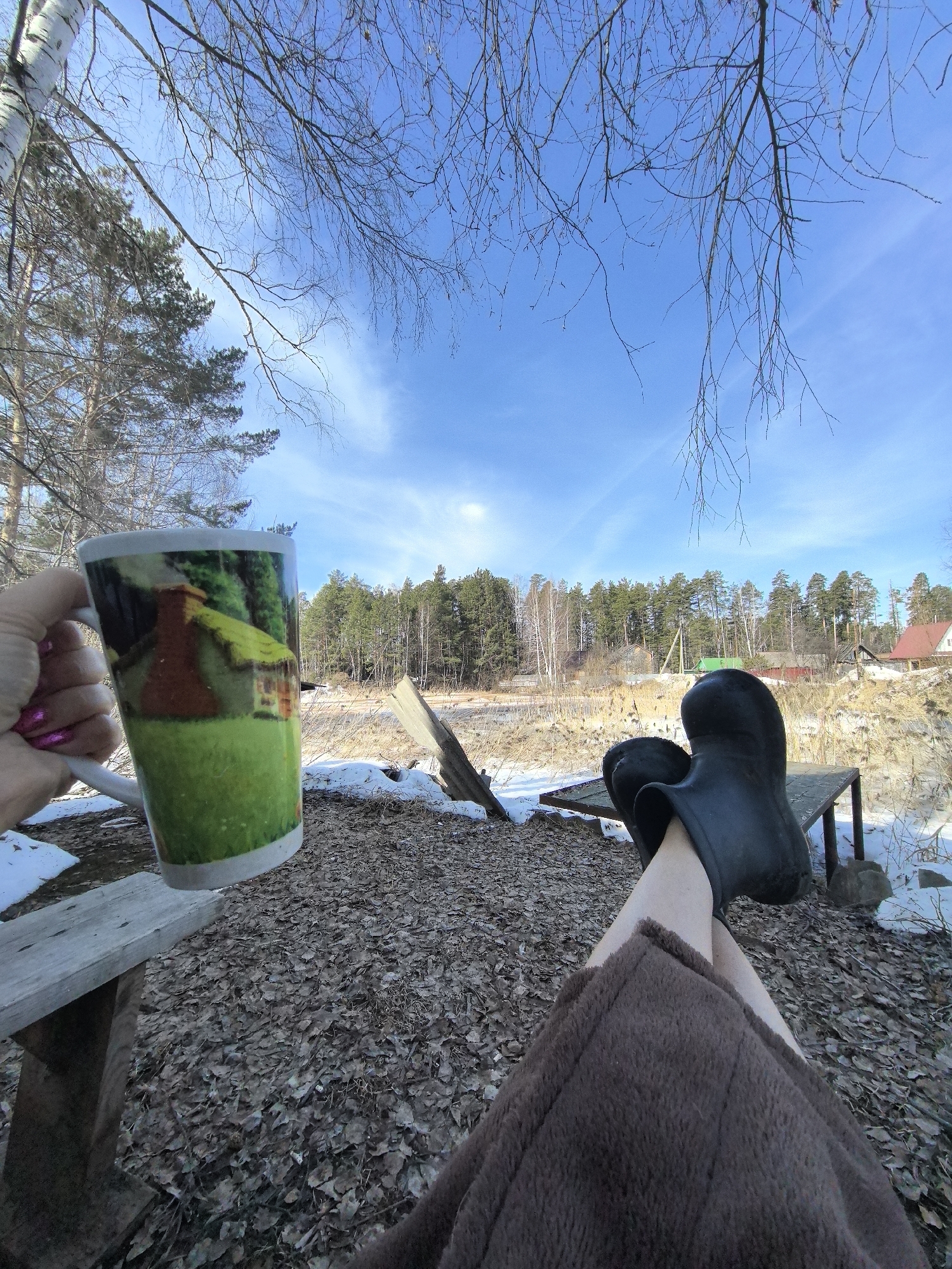 The image depicts a person relaxing outdoors, possibly in the vicinity of Noviy, Russia, suggested by the landscape and vegetation. The person is seated, legs extended onto a low table, wearing black galoshes and covered with a brown blanket. The setting appears to be a semi-wooded area with remnants of snow on the ground, suggesting early spring. A mug is held in one hand, displaying a pastoral scene on its surface. The background features a view of a flat area with sparse, dry vegetation and a line of trees. A few buildings are visible in the distance. The sky is blue with scattered clouds, adding to the outdoor ambiance.