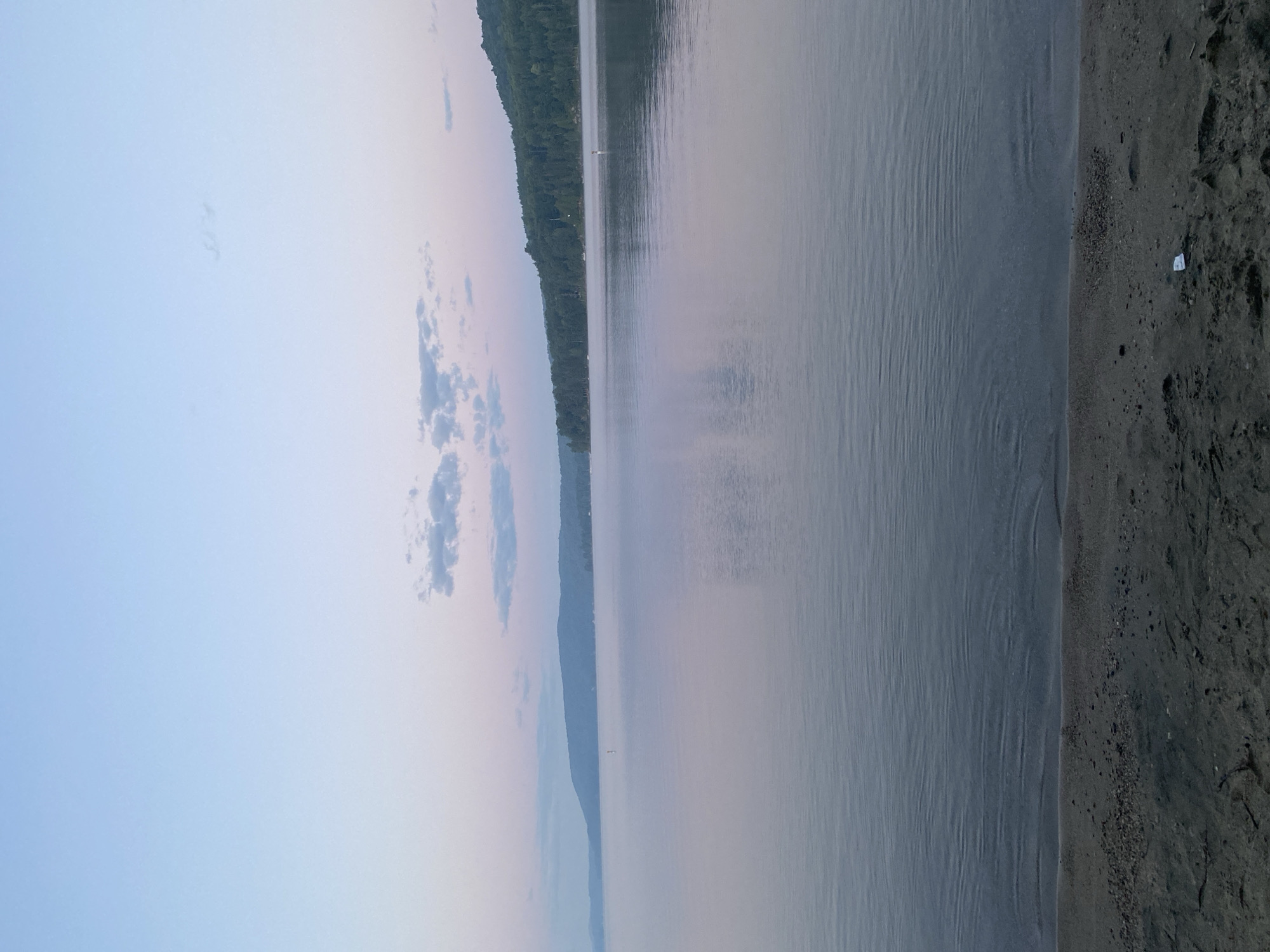 An atmospheric long-shot photograph captures a serene lake at dawn, with a light mist hovering over the water. In the distance, a tree-lined shore with rolling hills forms the backdrop. The sky transitions from a pale blue at the top to soft pink and lavender hues near the horizon, with scattered wispy clouds. In the foreground, a dark, sandy beach is visible, with small pebbles and sand grains. A white object, possibly a wrapper, lies on the sand near the water's edge. The scene suggests a tranquil natural environment, likely in Duhamel, Canada, given the presence of a lake and lush vegetation.